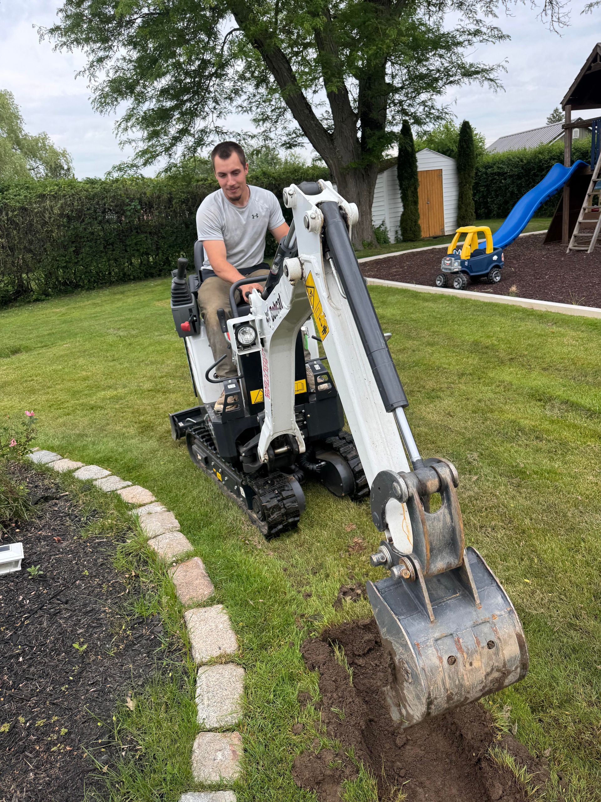 A person operates a mini-excavator to dig a trench in a grassy backyard, near a flower bed and a playground.