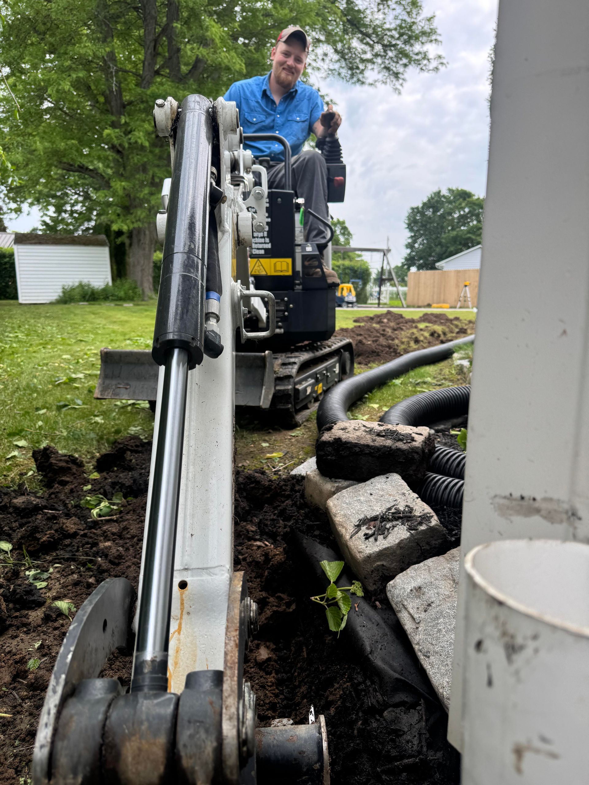 Man operating a mini excavator, digging a trench near a building.