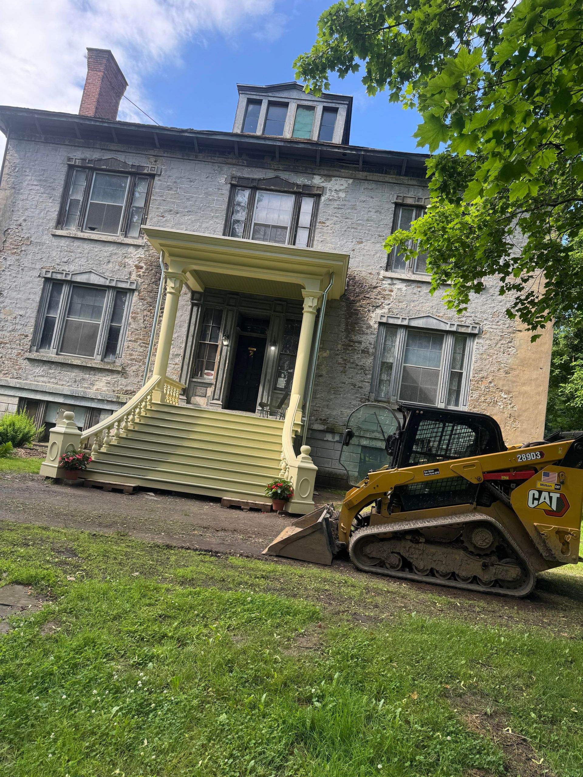 Skid steer loader in front of a dilapidated multi-story building with a porch and steps.