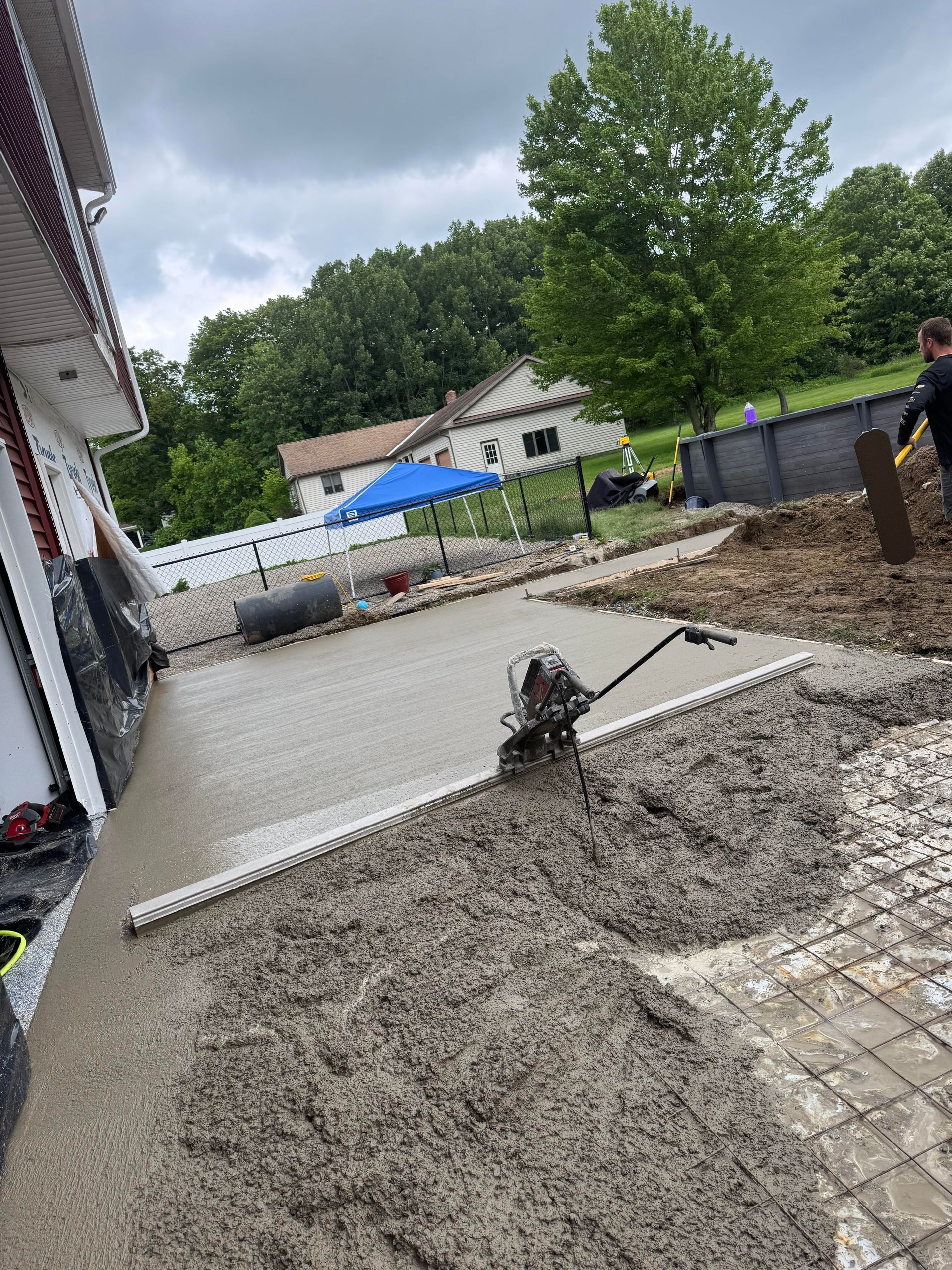 Freshly poured concrete patio being leveled, with person and tools, near a house with trees in the background.