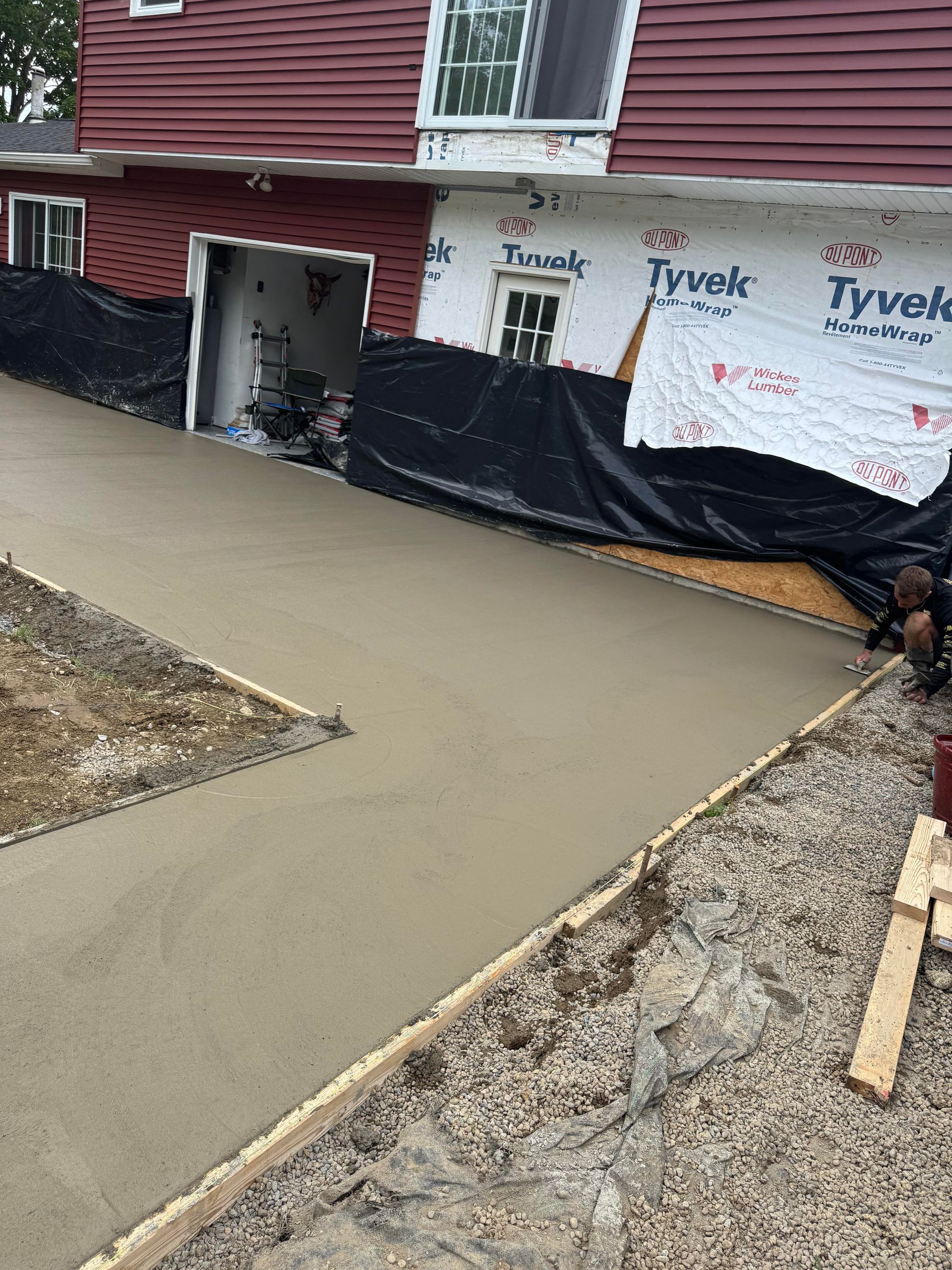 Freshly poured concrete driveway leading to a house under construction. Brown siding, red roof, Tyvek wrap visible.