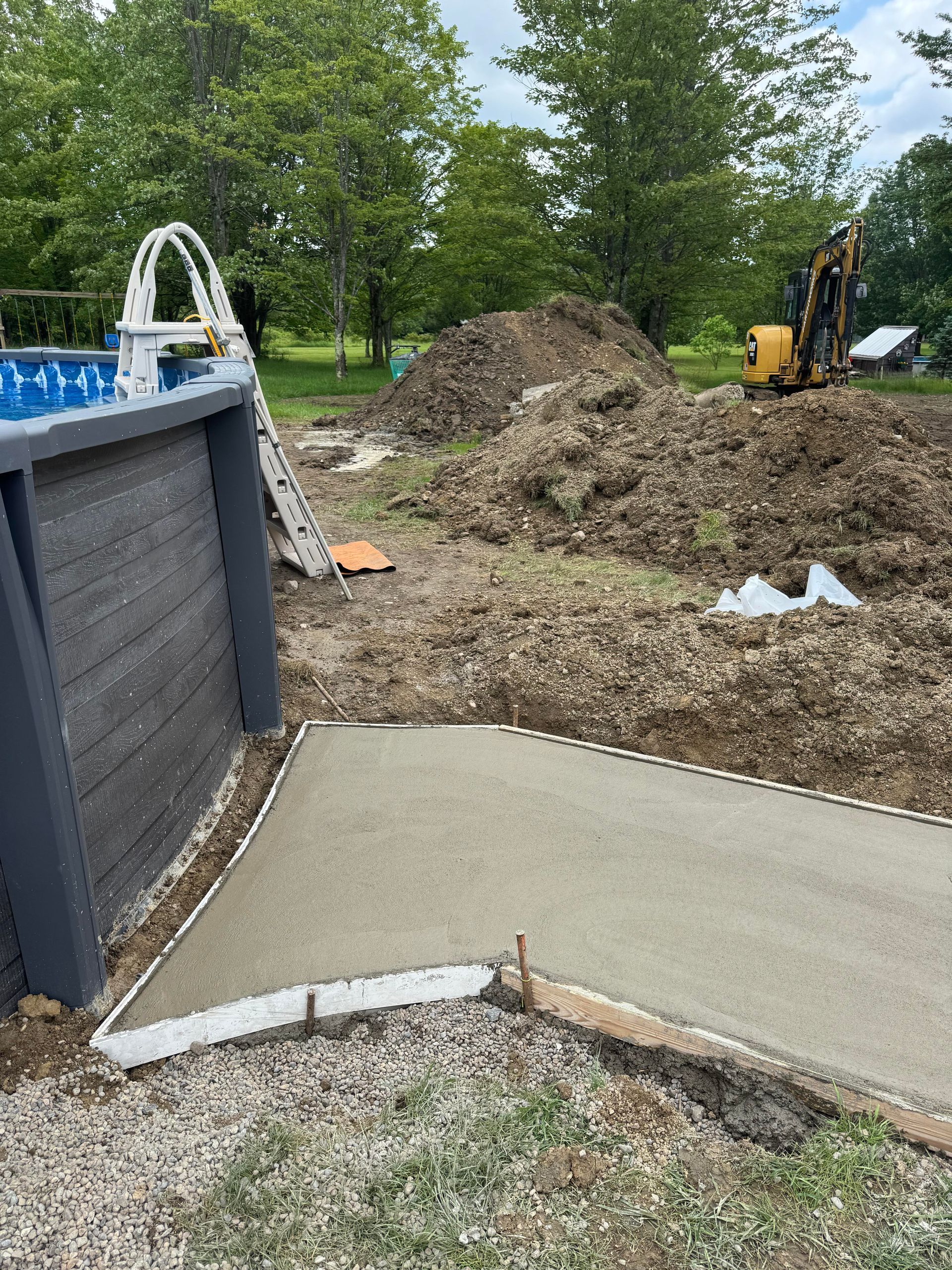 Concrete patio under construction next to an above-ground pool, with a pile of dirt and a small excavator.