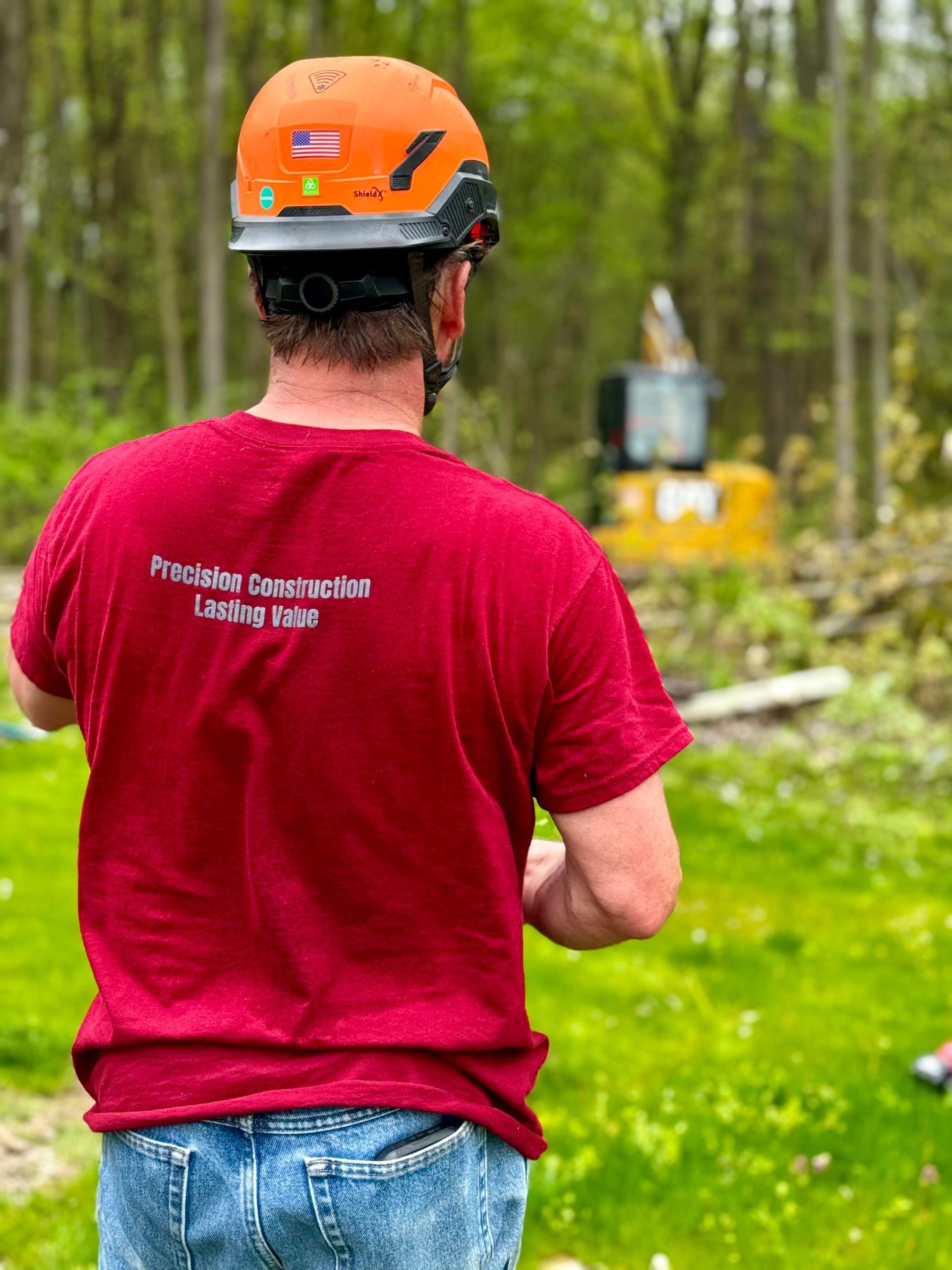 Man in orange hard hat, red shirt, and jeans surveys a construction site with an excavator in the background.