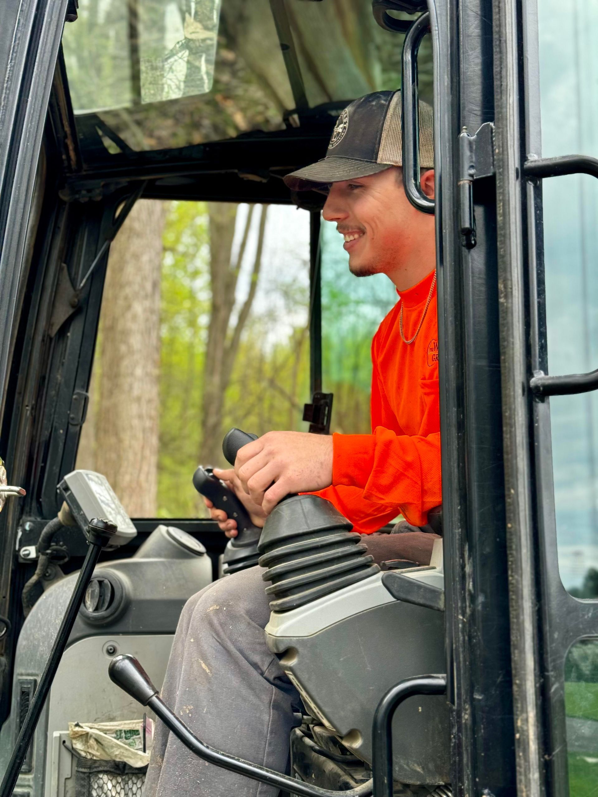 Man operating machinery, smiling. Inside cab, wearing orange shirt and hat. Trees in background.
