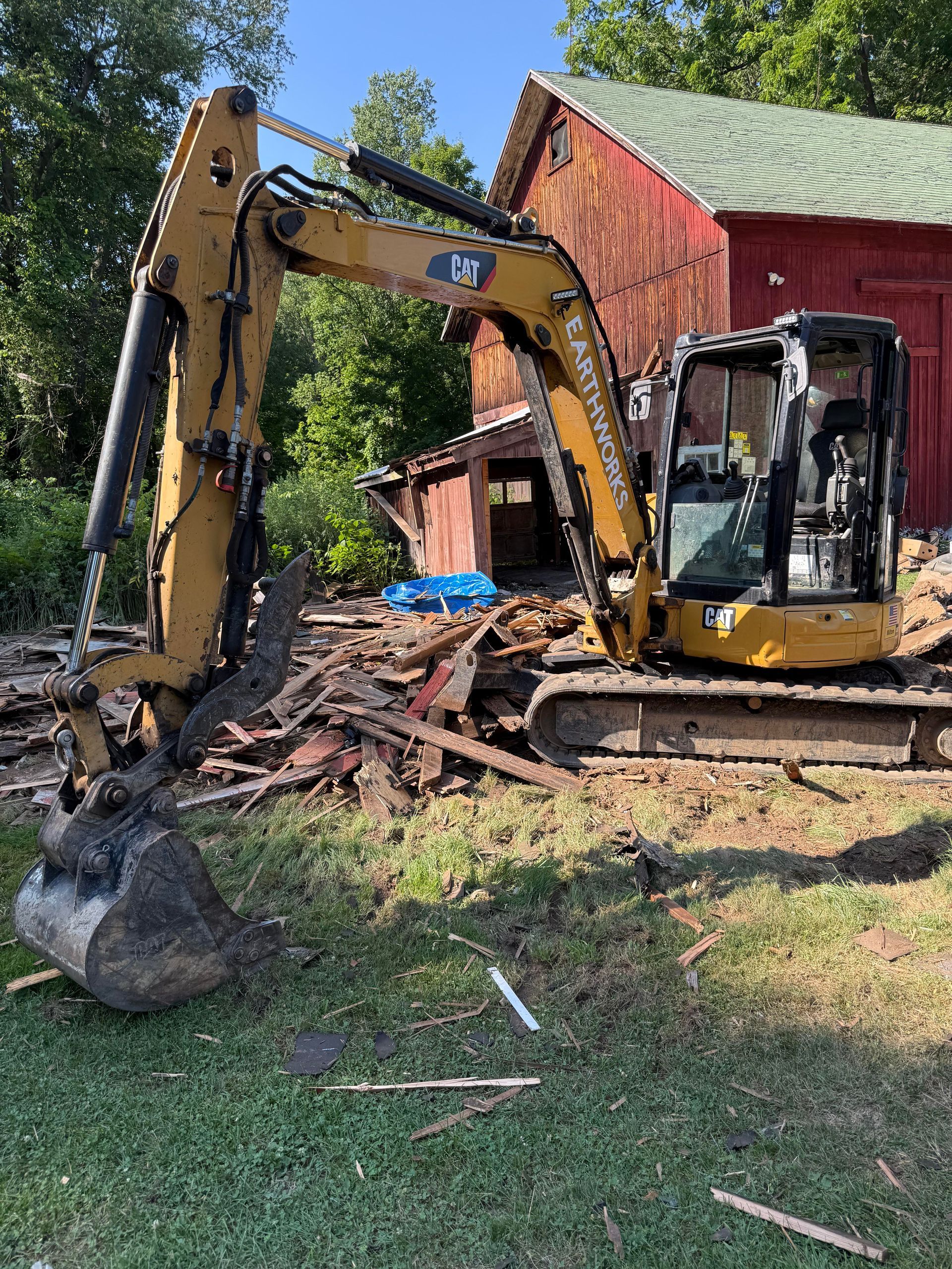 Yellow excavator demolishing a red barn, debris scattered on the ground. Green grass and trees in background.