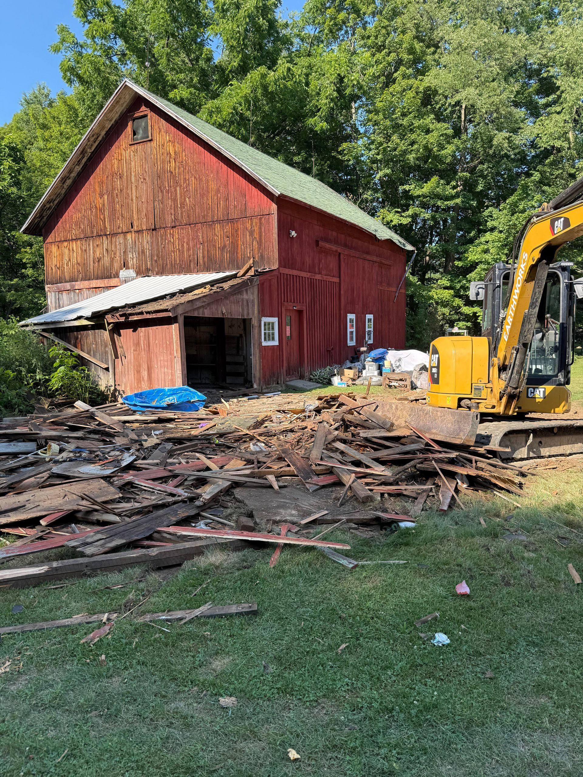 Red barn with a damaged lean-to, debris in the foreground, and an excavator.