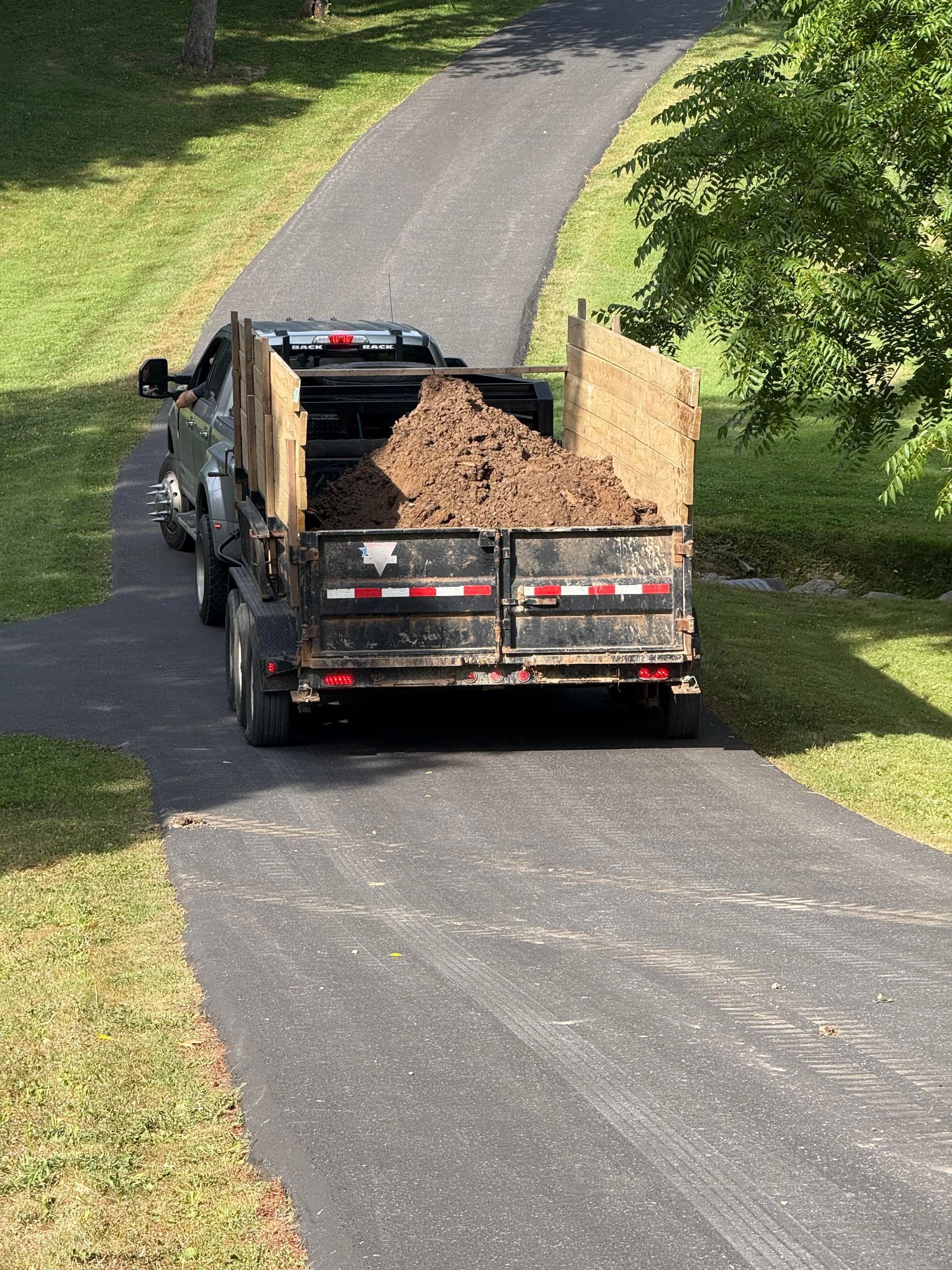 Truck bed filled with dirt on a black asphalt driveway, surrounded by green grass and trees.