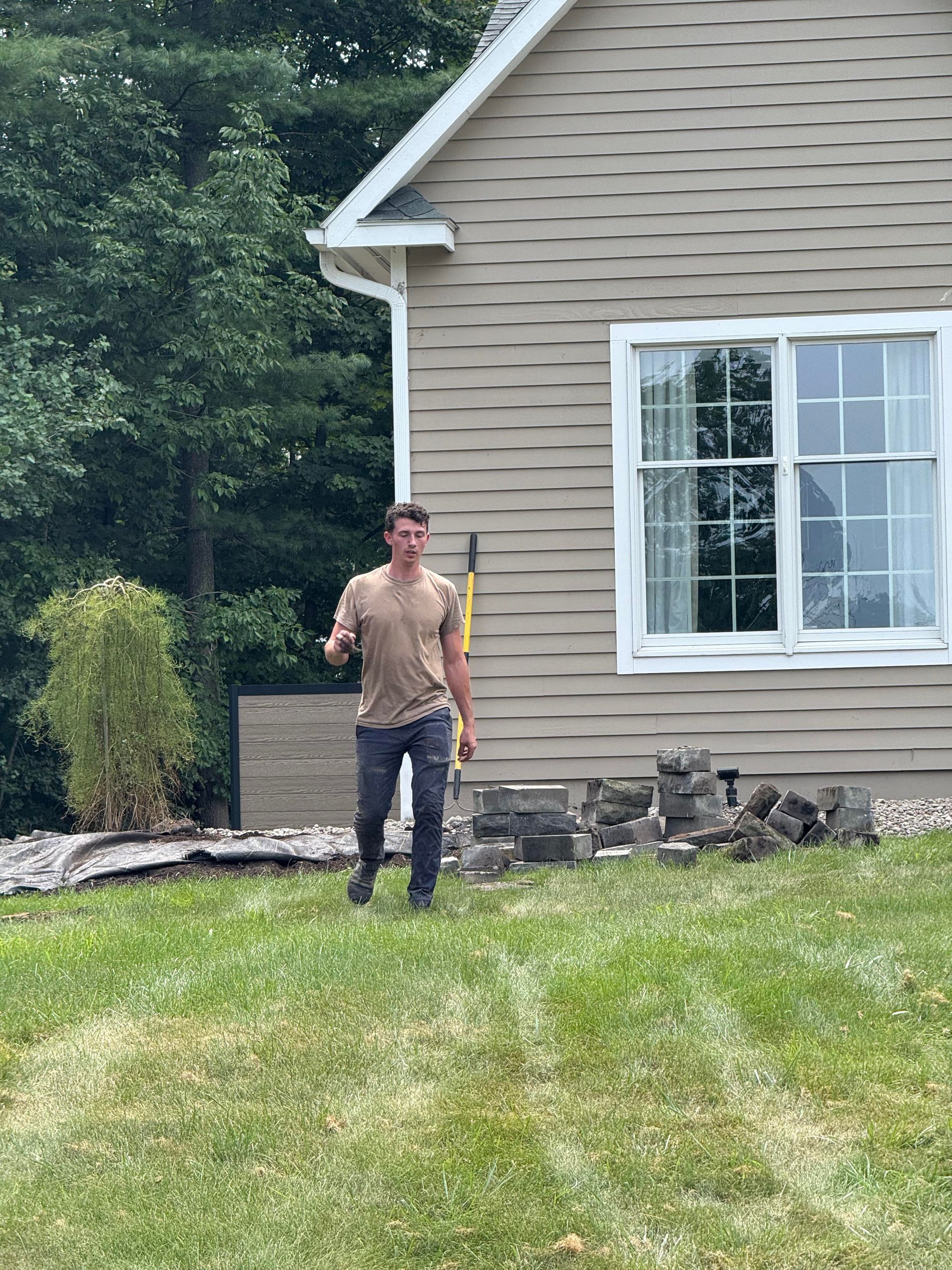 Man walking on grass near house, gesturing with hand, next to bricks and shovel.