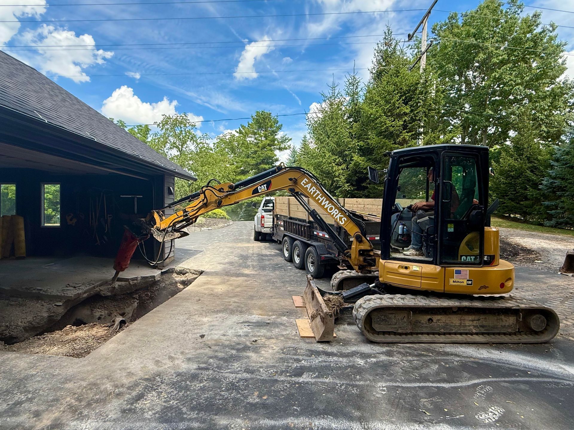 Yellow excavator breaking up concrete near a building on a sunny day. A trailer is nearby.