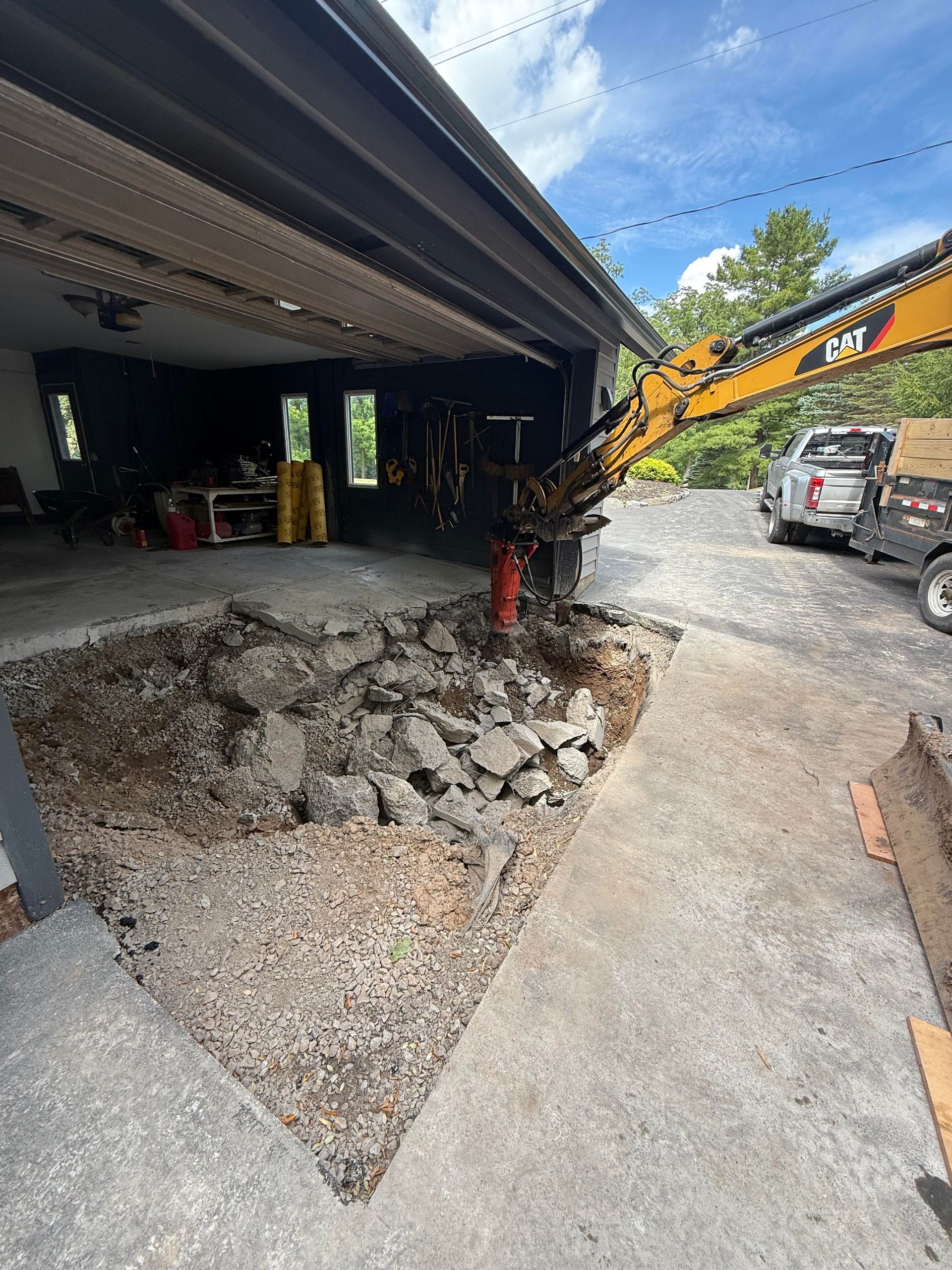 Construction site: excavator tearing up concrete driveway next to garage.