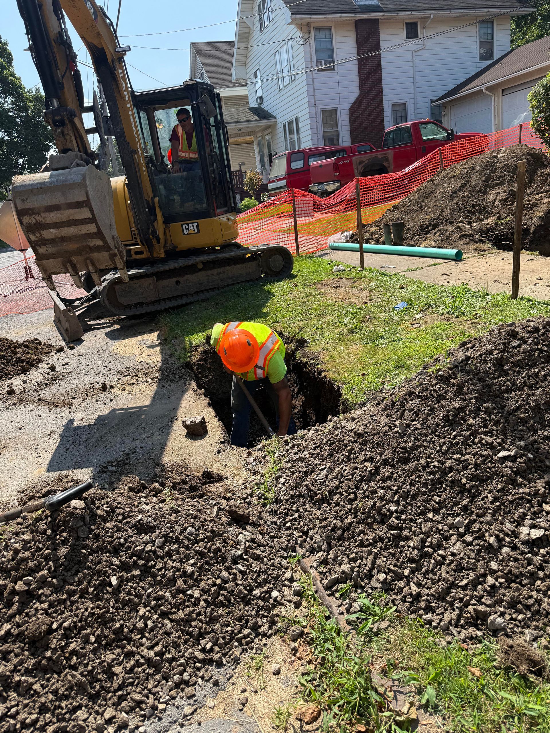 Construction worker in orange vest working in a hole, excavator nearby, orange safety fencing, residential street.