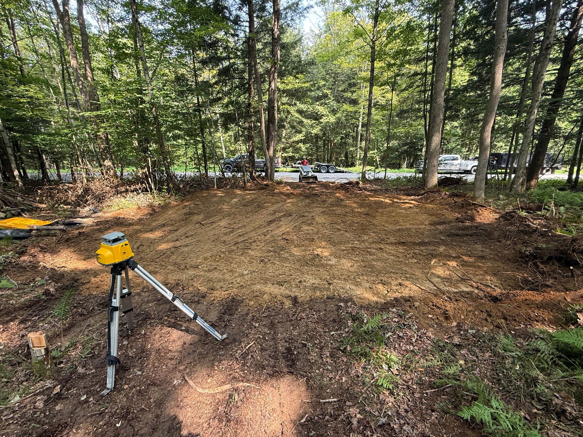 A leveled dirt area in a wooded area; a yellow laser level on a tripod in the foreground.