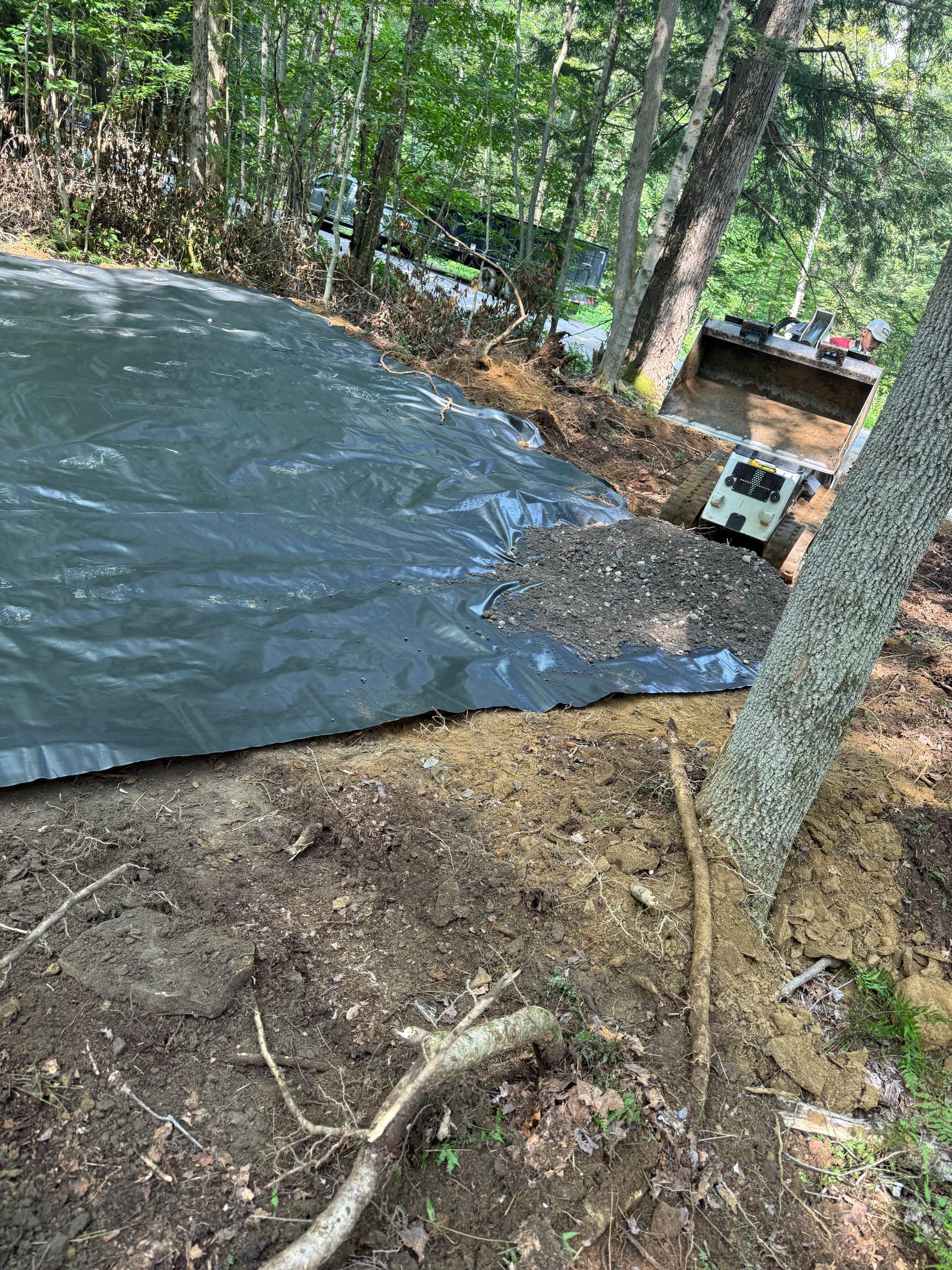 Black landscape fabric covers a sloped area, gravel and dirt around a tree, with a vehicle in the background.
