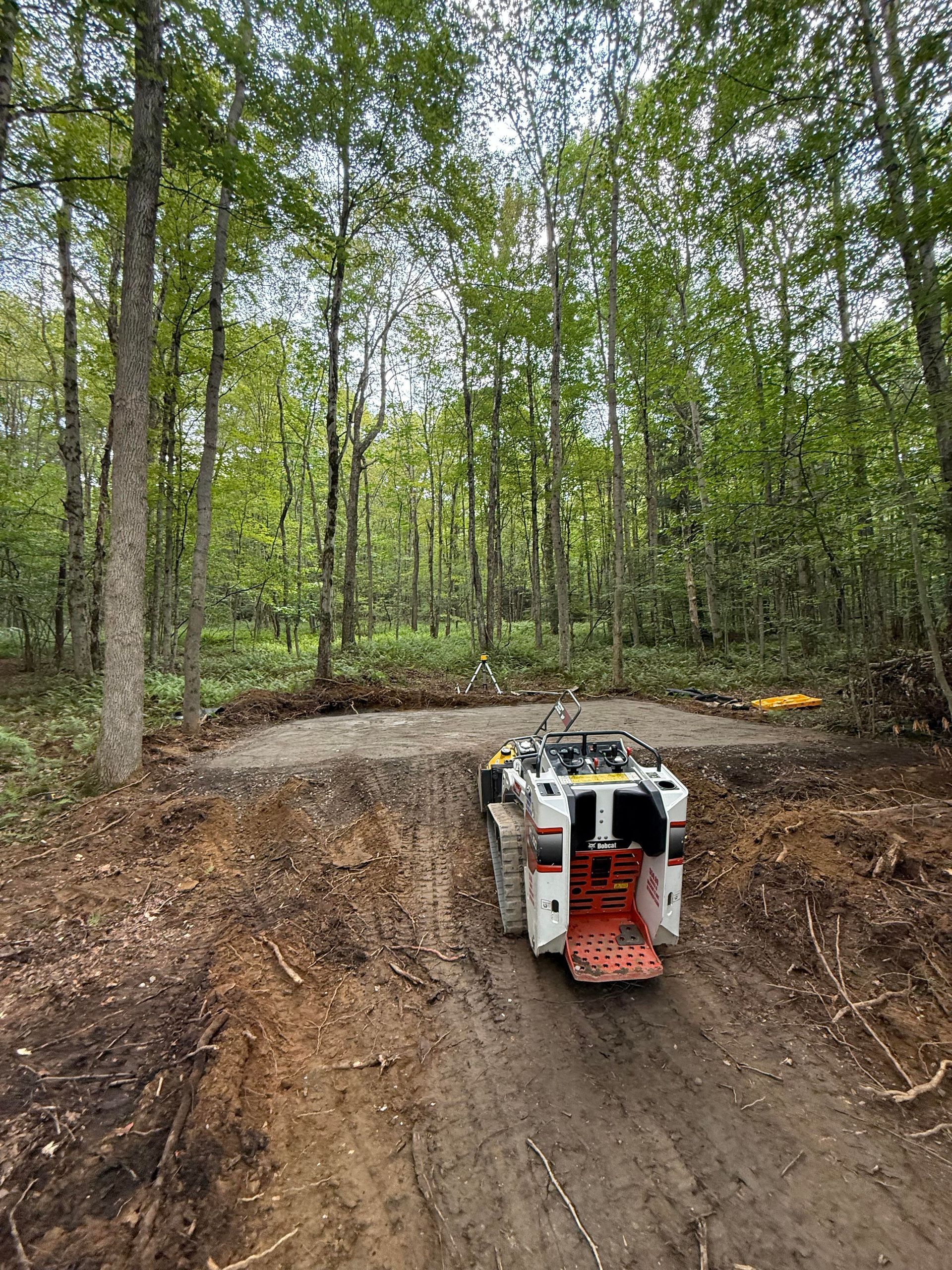 A small, white construction vehicle on a dirt path in a forest clearing, surrounded by trees.