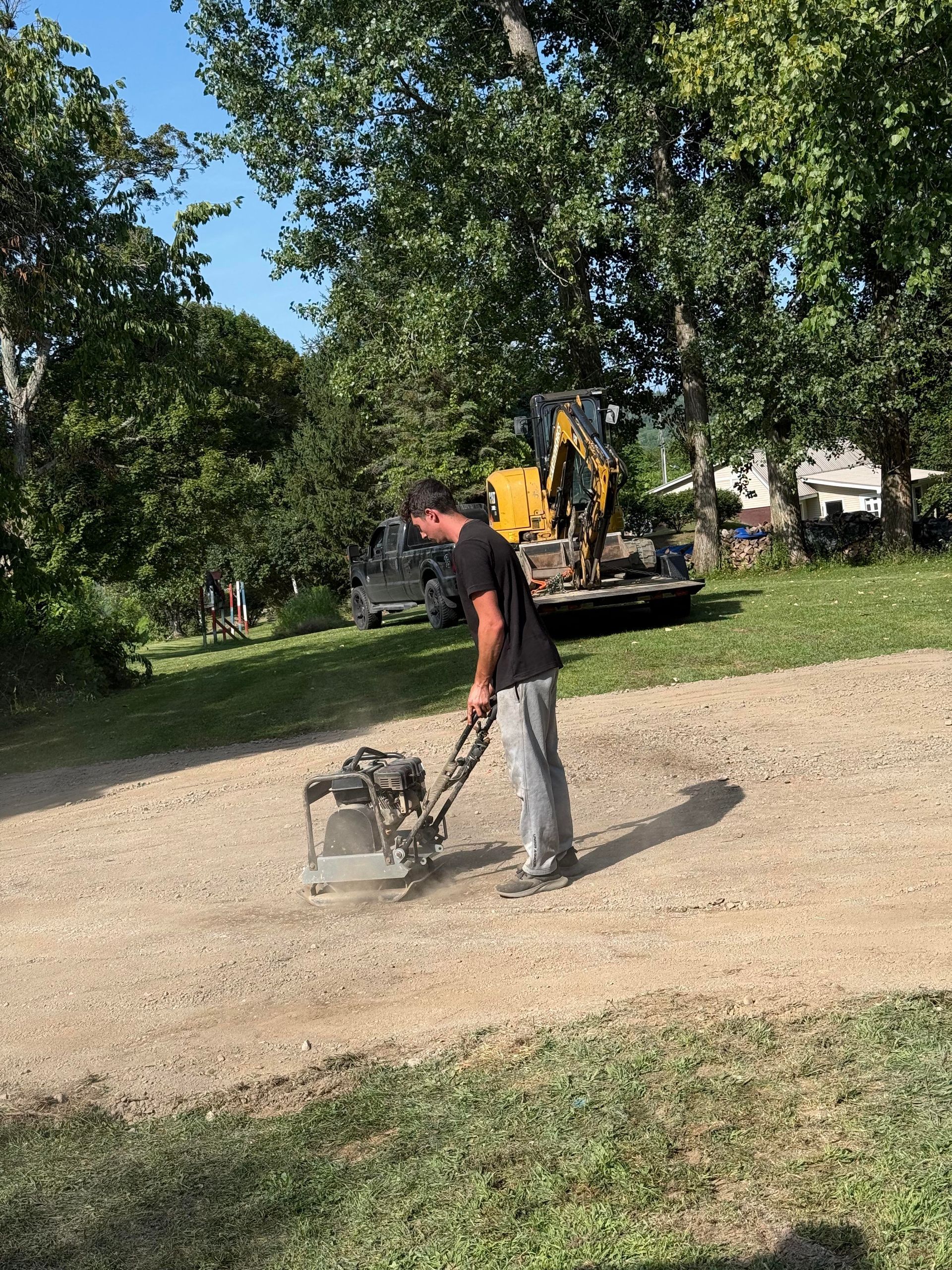 Man compacting gravel driveway with a vibratory plate compactor; yellow excavator in the background.