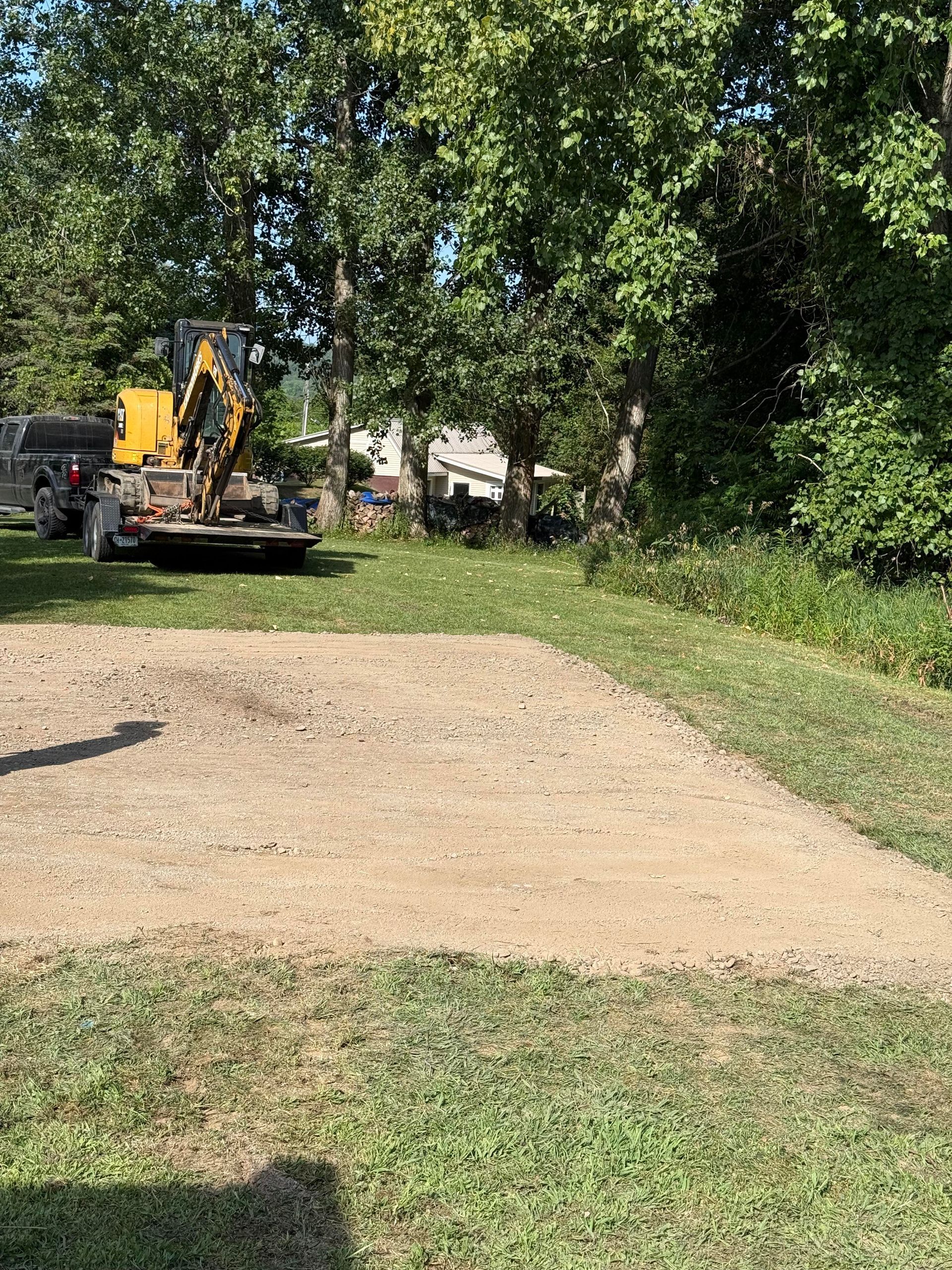 Gravel patch on grass; small excavator in the background. Trees in background, sunny day.