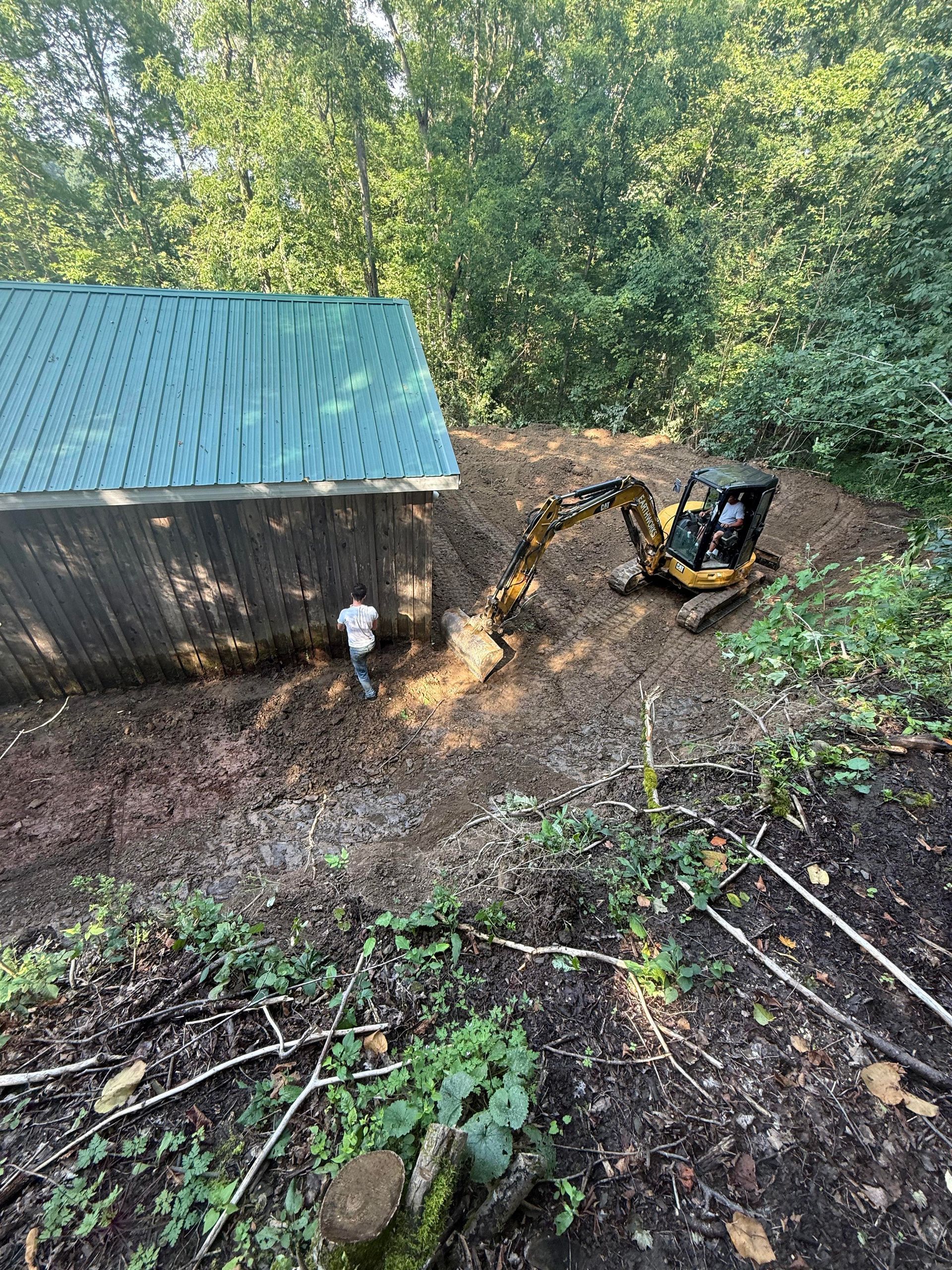 Excavator digging near a wooden structure with a green roof; a person stands nearby. Forest setting.