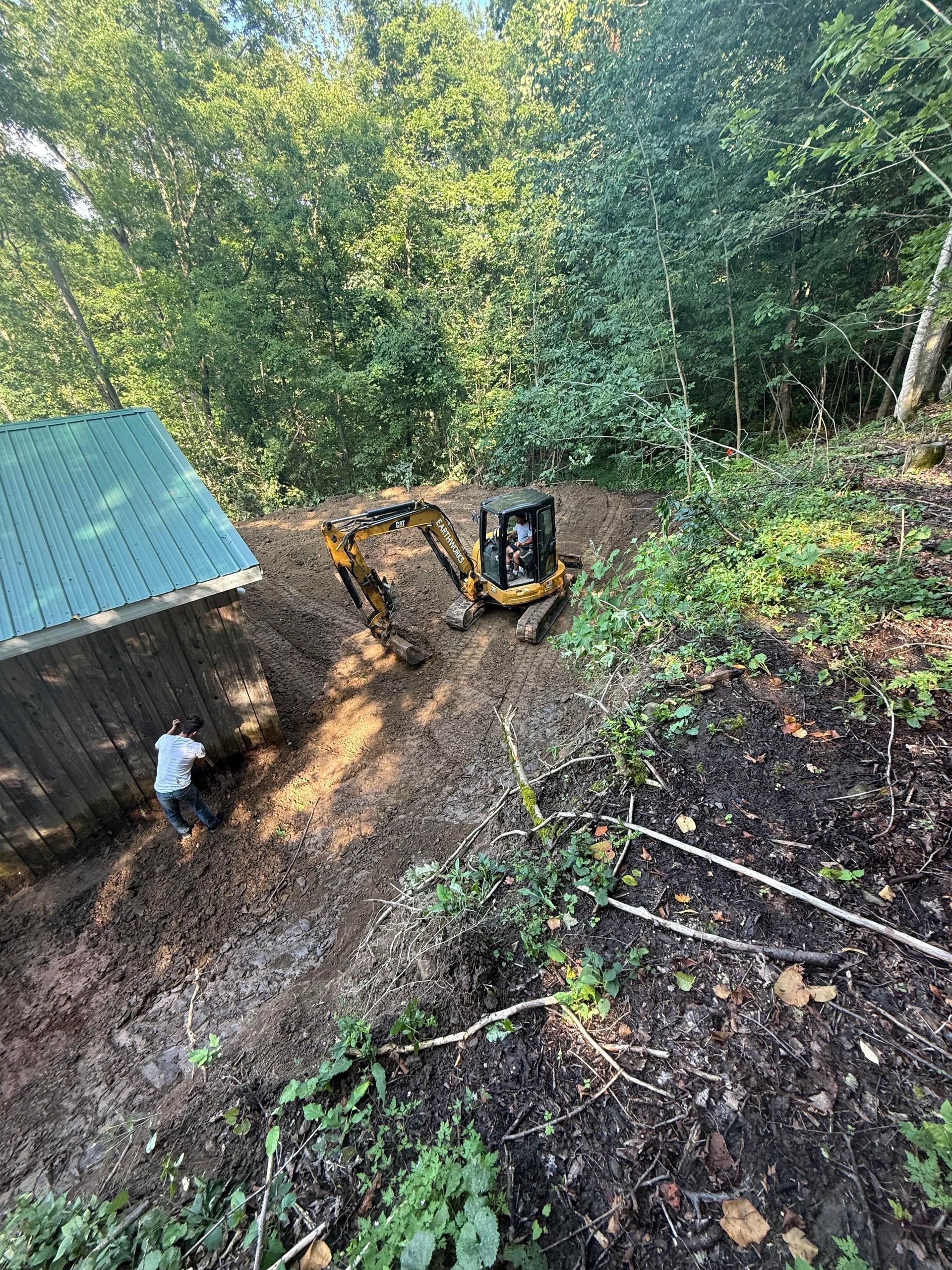 An excavator on a wooded slope clearing land near a building. A person is near the building.
