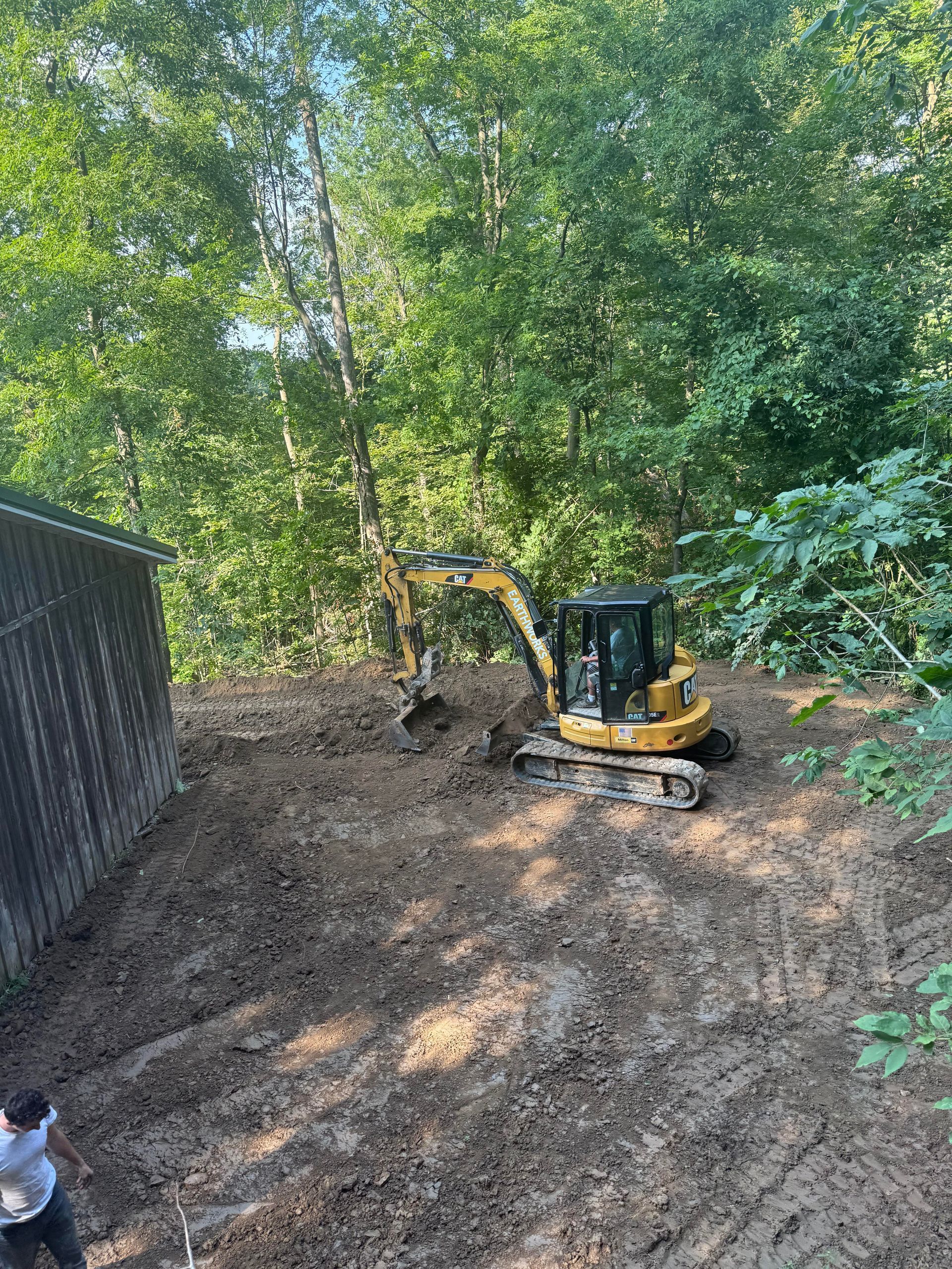 Yellow excavator on a muddy hillside near a wooden structure and trees. A person stands near the structure.