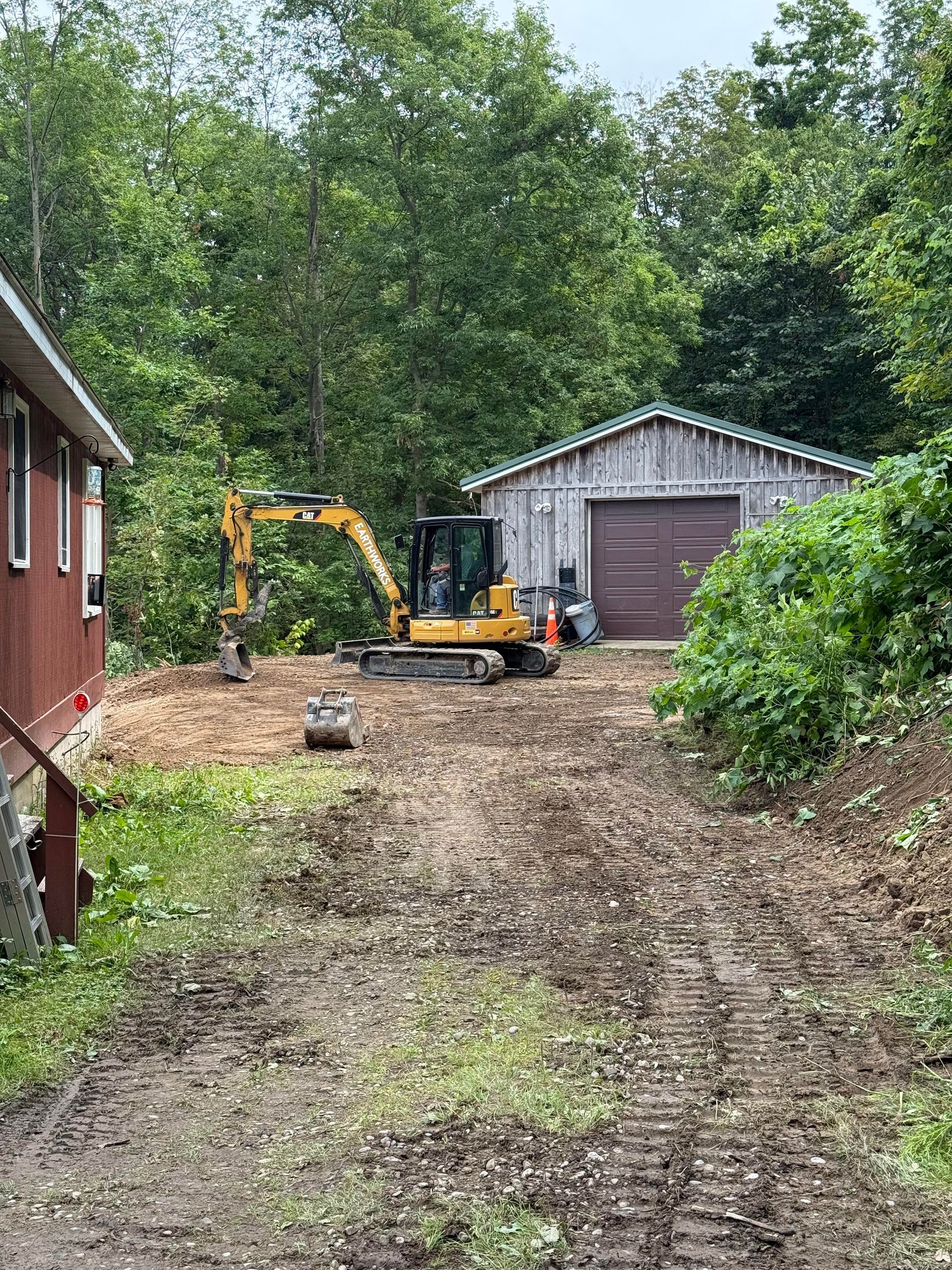 Yellow excavator on a dirt driveway clearing land near a red building and a gray garage.