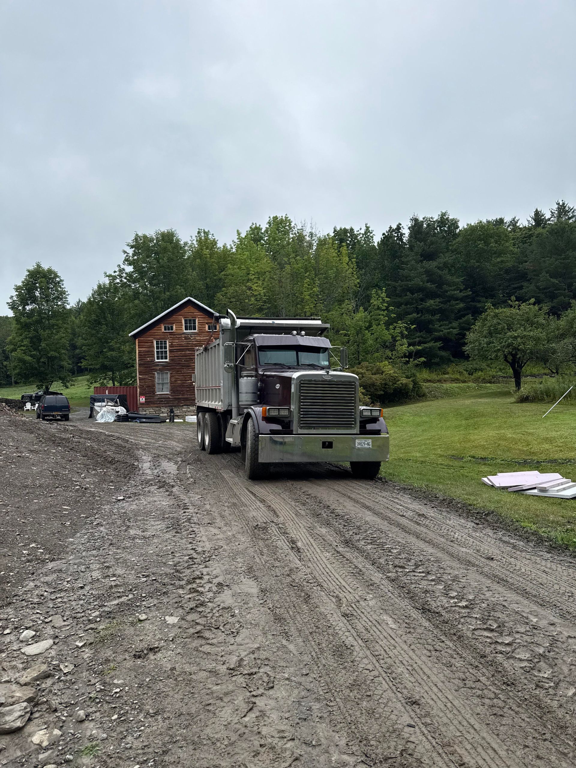 Muddy road with a dump truck in front of a small house and trees. Overcast sky.