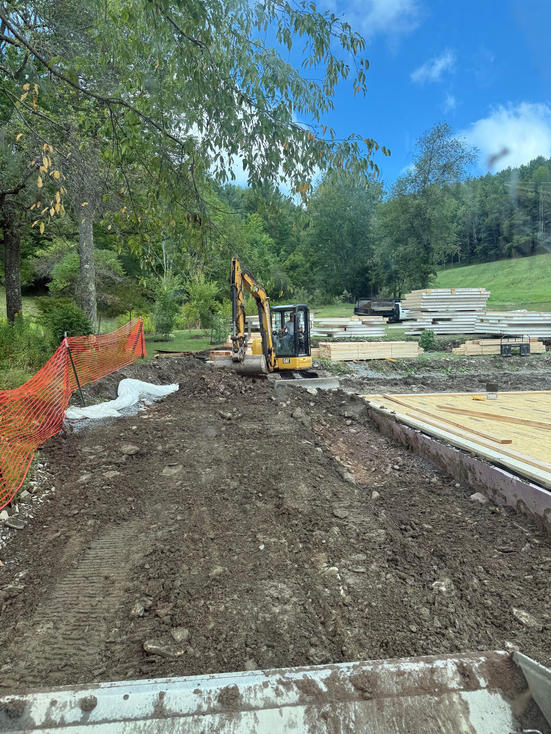 Excavator digs in a dirt lot. Orange fencing, lumber, and trees are in the background. Bright sky.