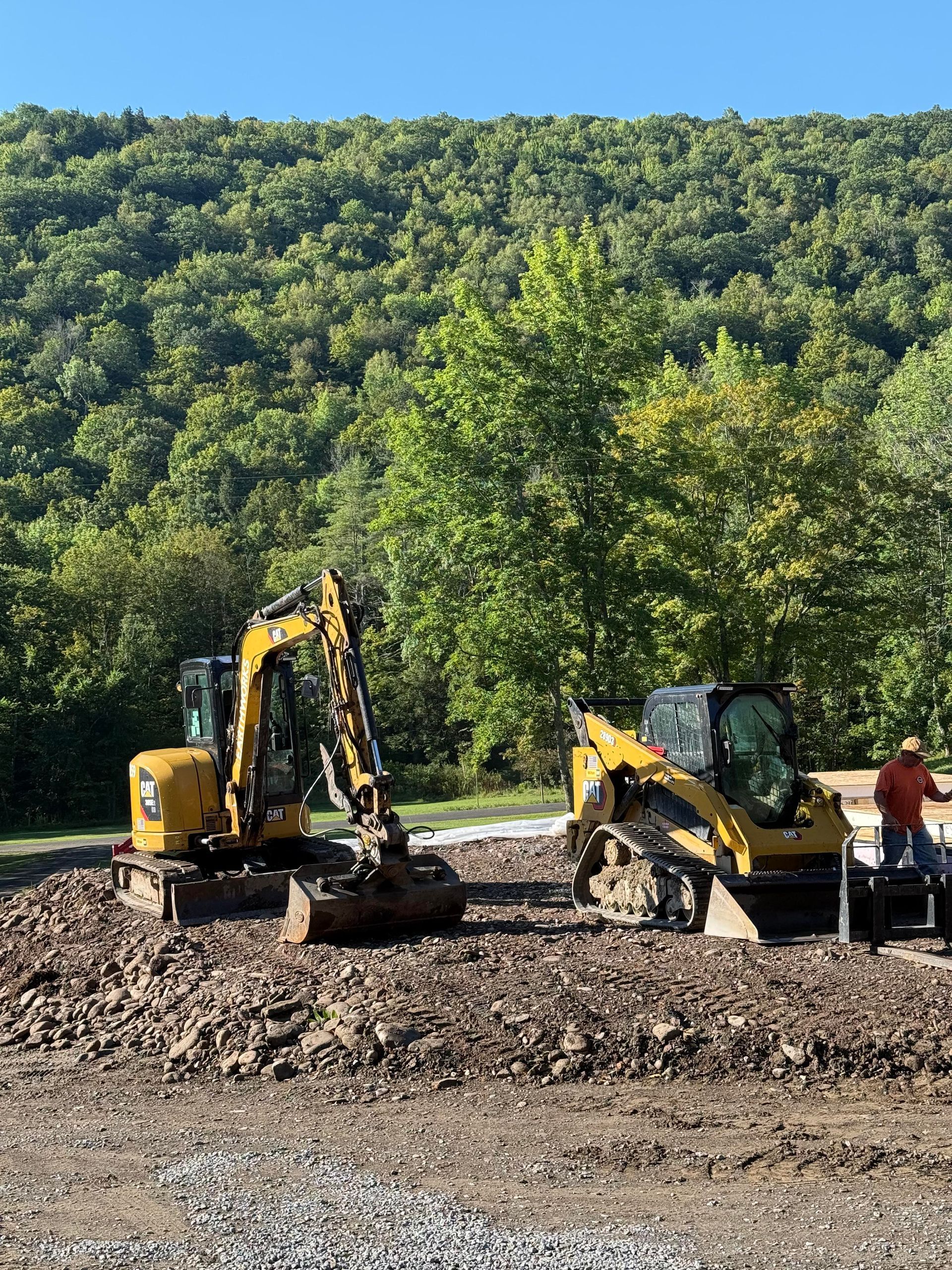 Two yellow construction vehicles on a dirt patch, forest background.