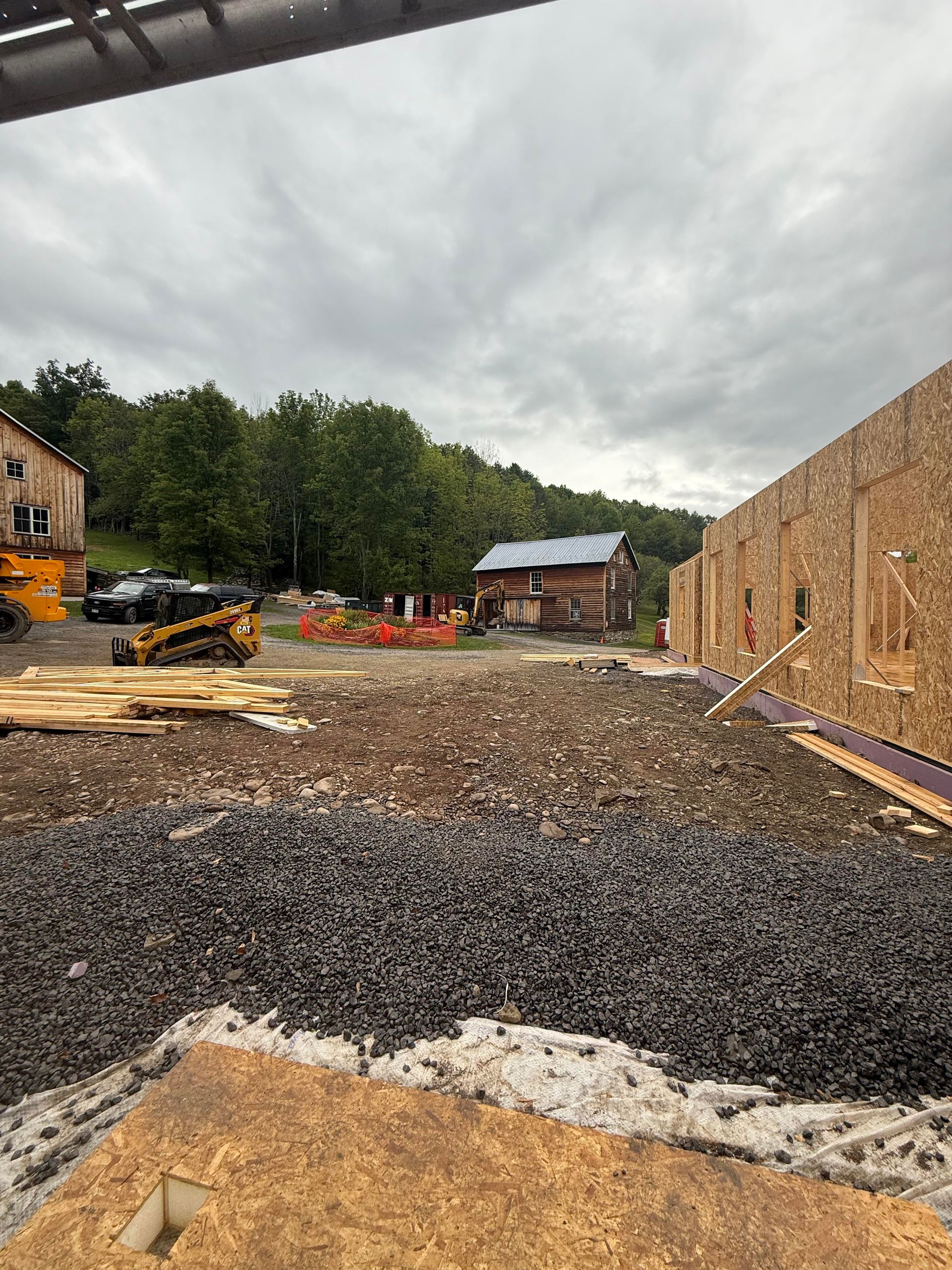 Construction site with a partially built structure, gravel, a small log cabin, and heavy machinery under an overcast sky.