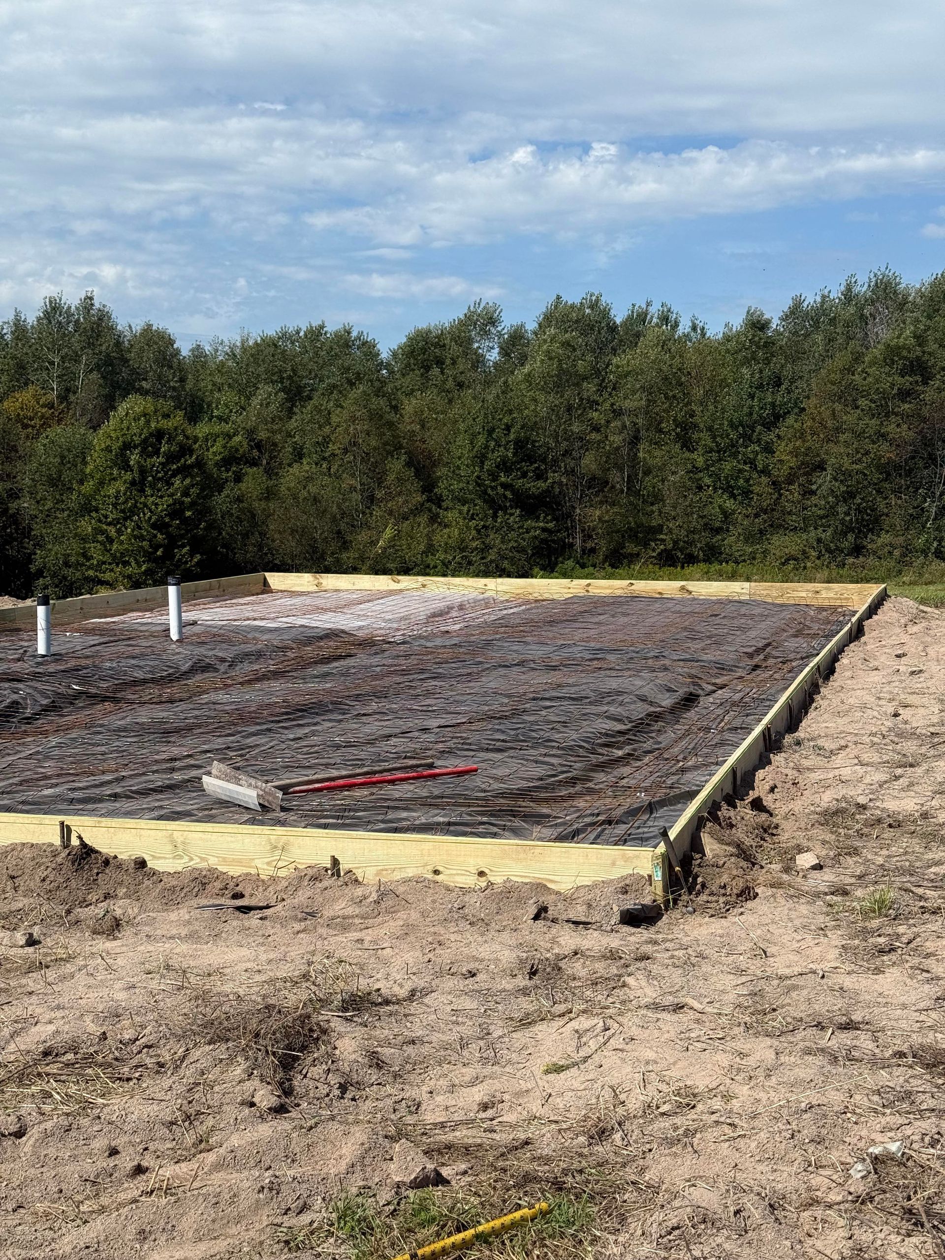 Construction site: wooden forms, rebar, and pipes outline a circular foundation, brown dirt, trees, sky.