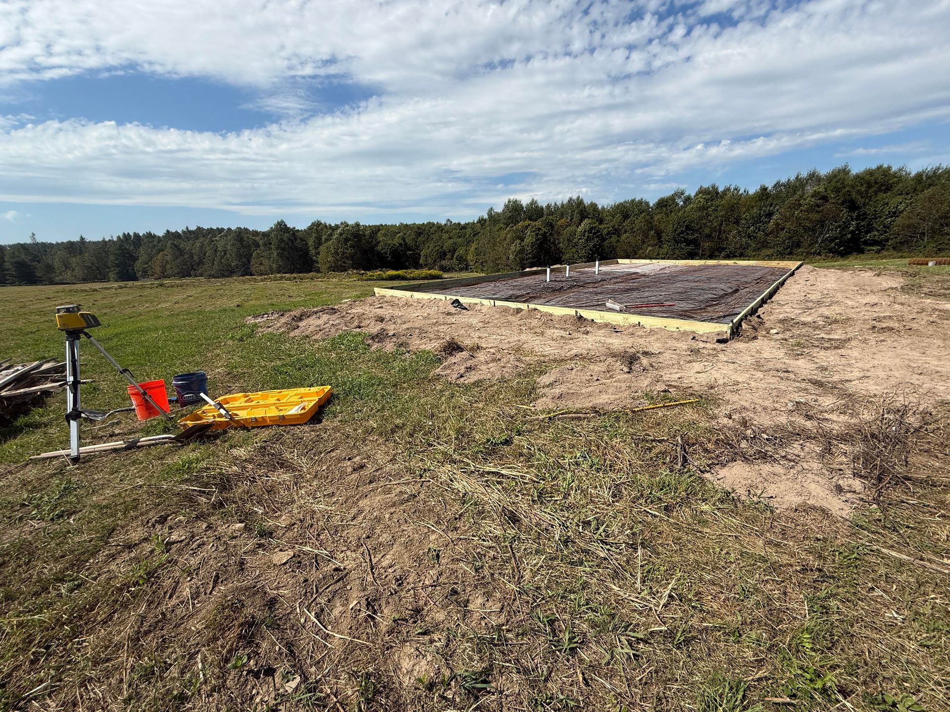 Open field with surveying equipment, a rectangular construction area, and forest in the background under a blue sky.