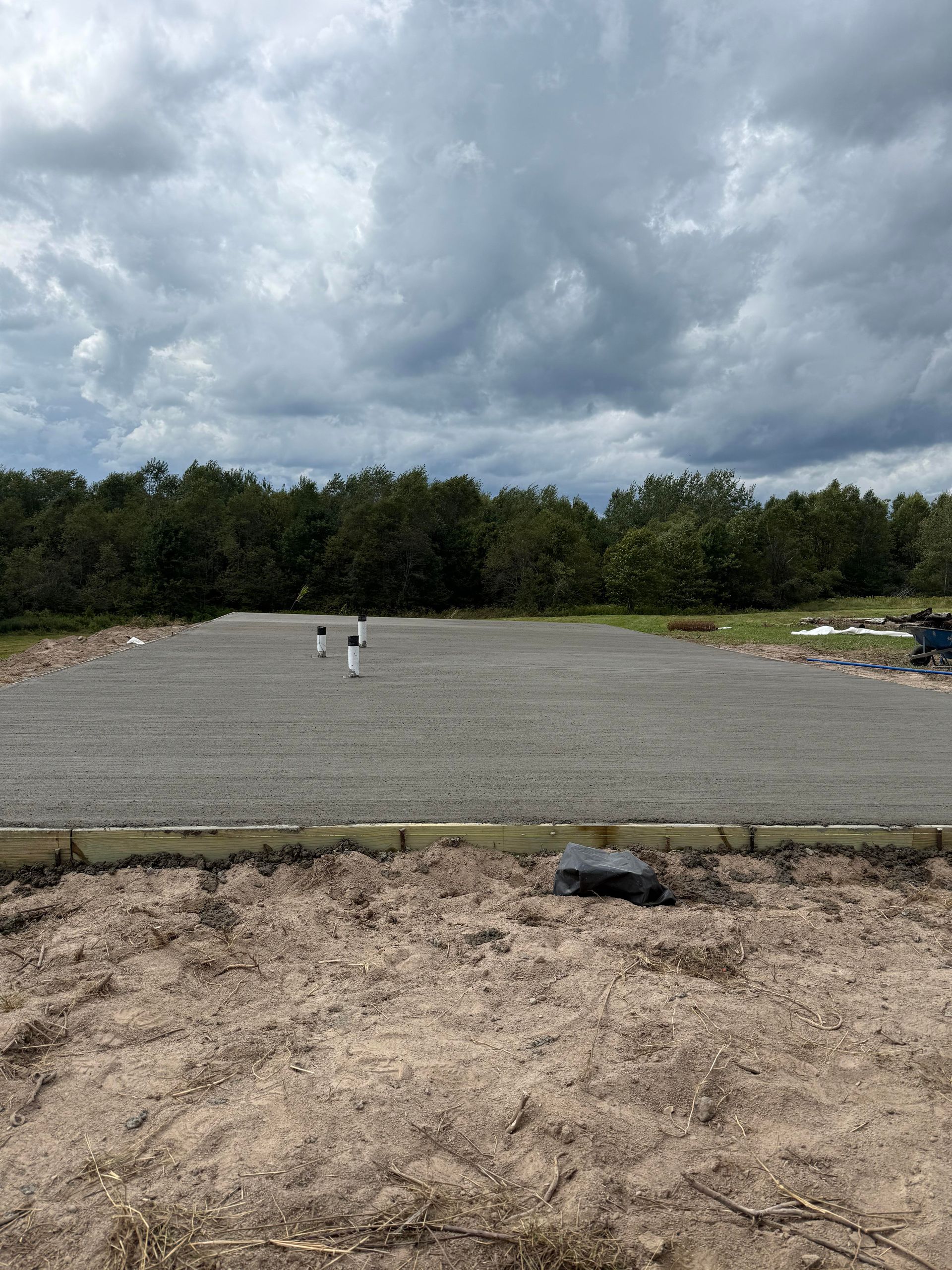 Gravel-covered lot with white pipes, dirt foreground, treeline background, cloudy sky.