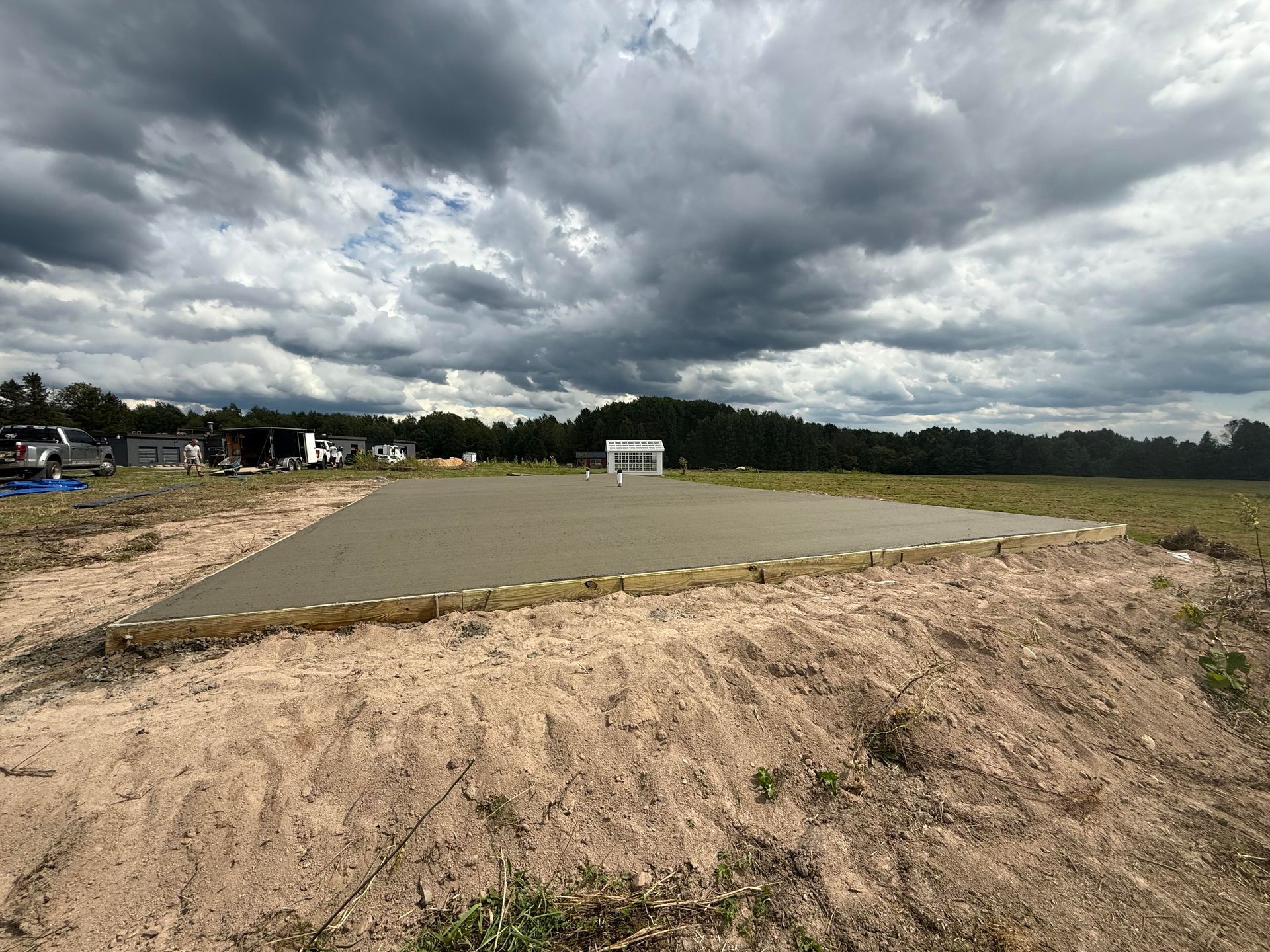 Newly poured concrete foundation in a field, surrounded by sand, wooden frame, and overcast sky.