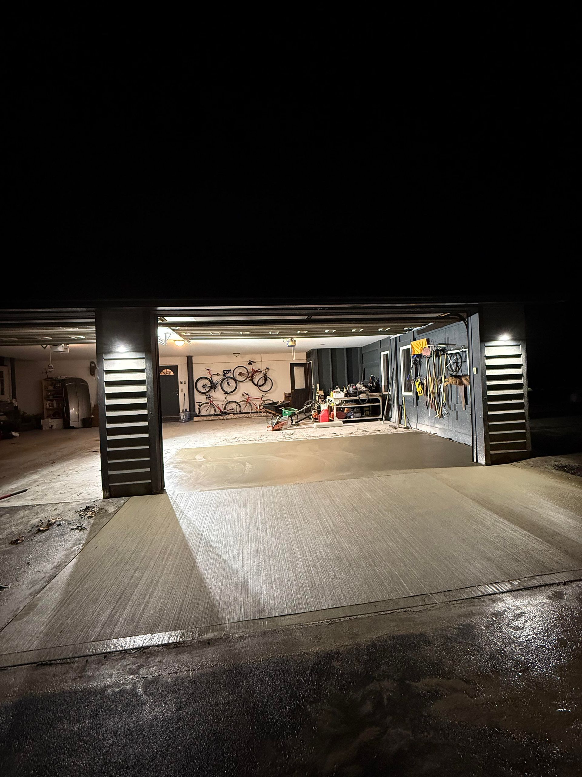 Garage at night with lights on, tools and a bicycle visible inside. Concrete driveway with tire tracks.