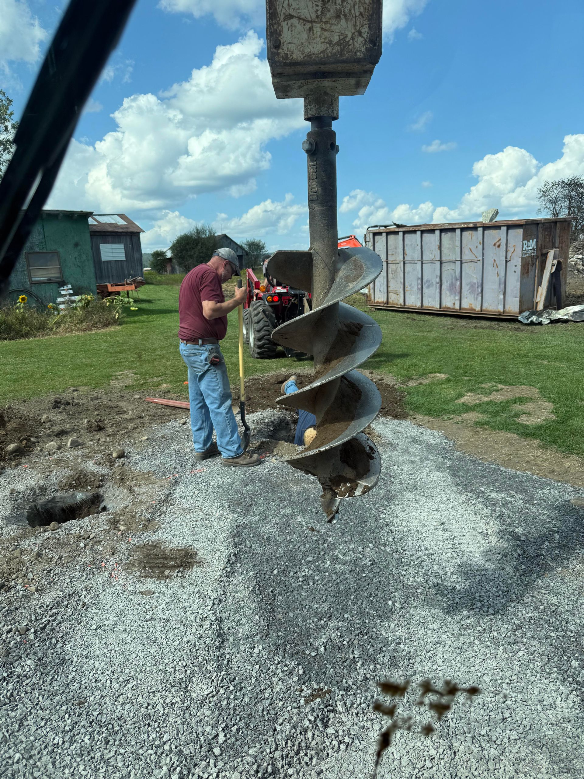 Man operating an auger drill, drilling into gravel, under a blue sky.