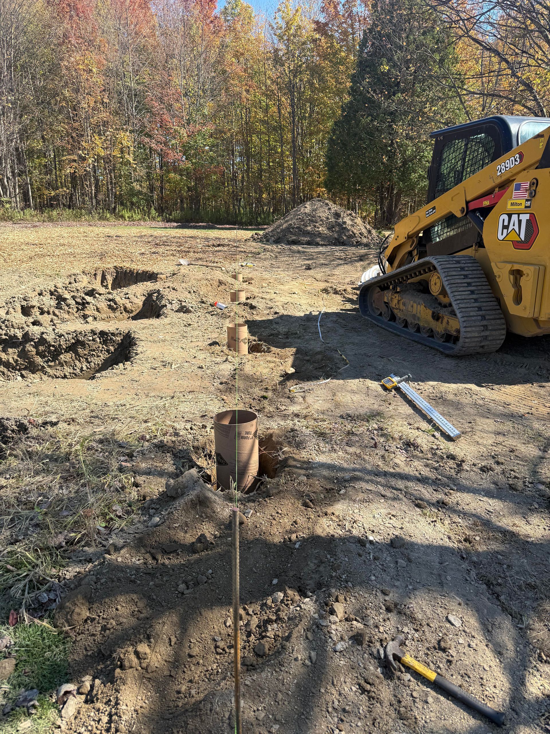 Construction site with a yellow Caterpillar skid steer, post holes, and a partly wooded background.