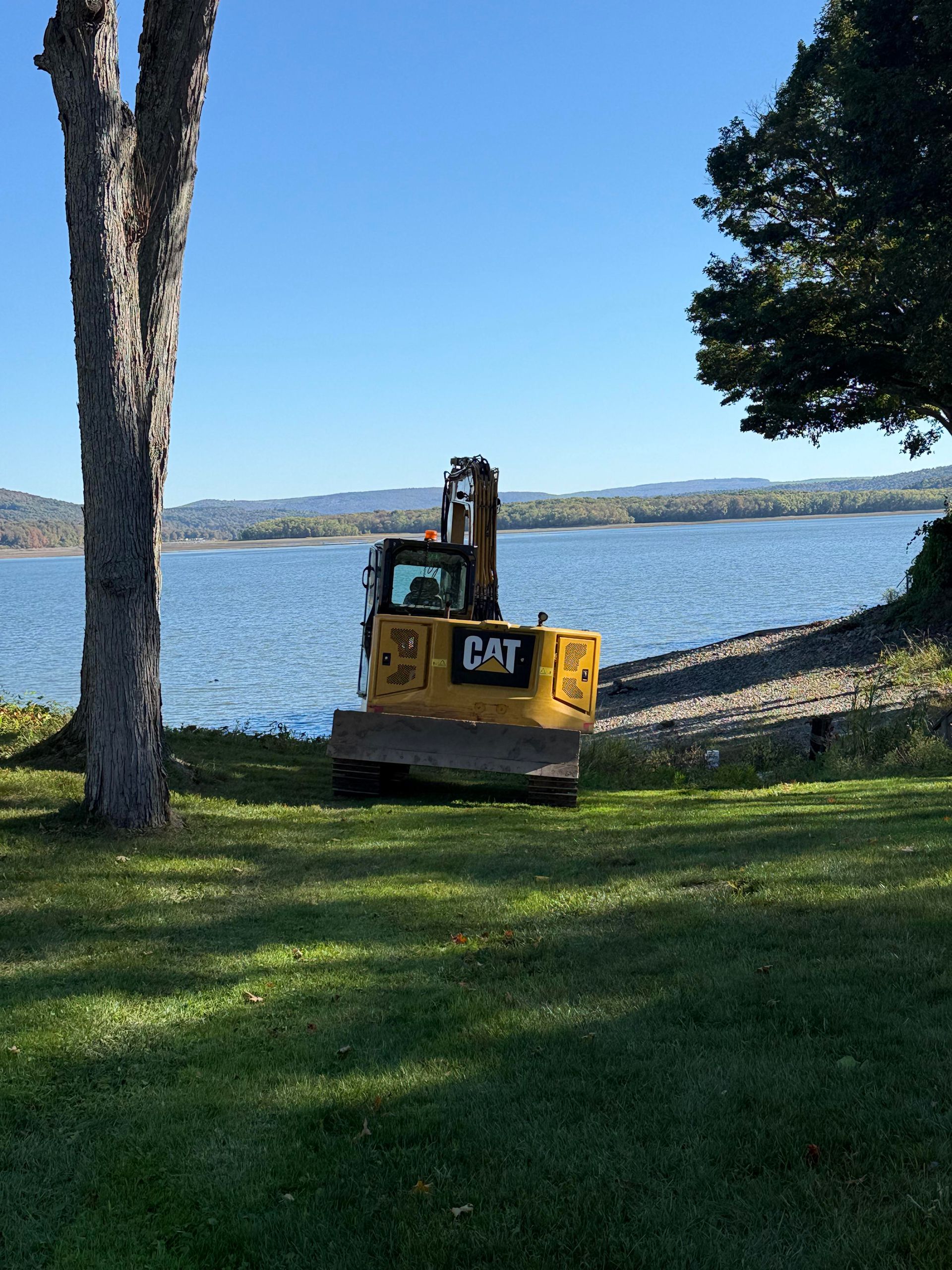 Yellow Caterpillar excavator on a grassy shore by a lake, under a bright blue sky.