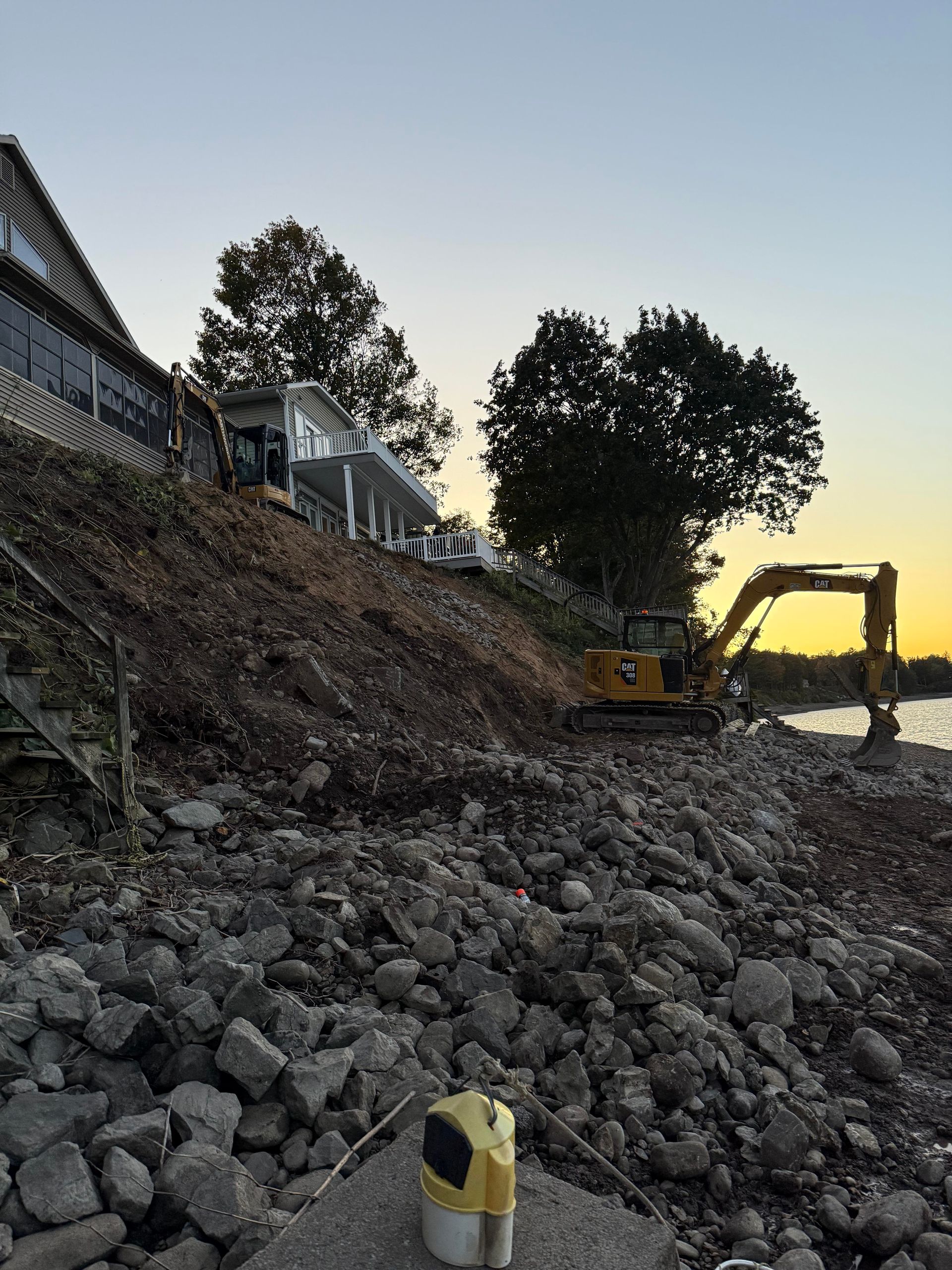 Excavator on a rocky shoreline, working near a house at dusk.
