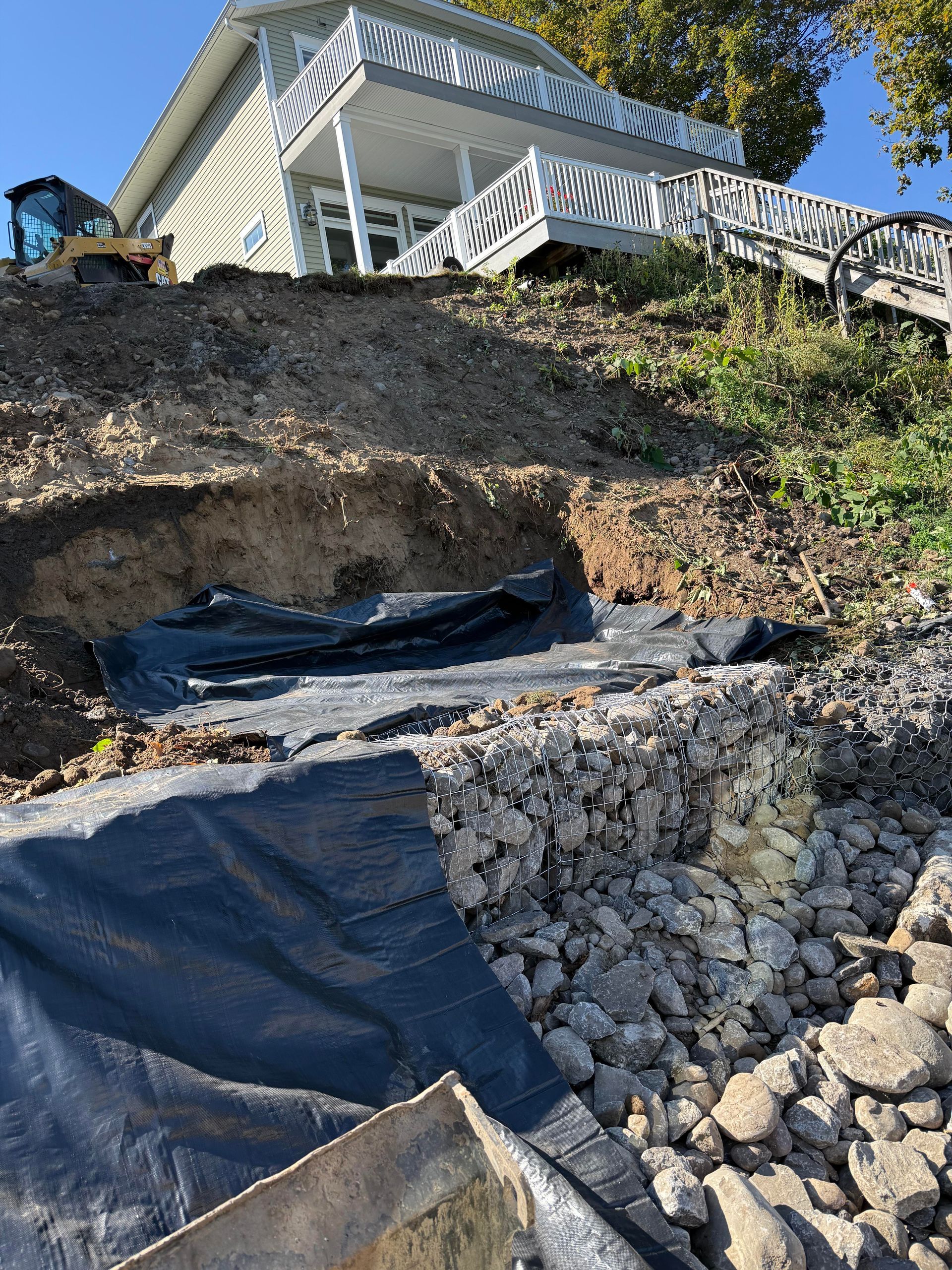 Construction on a hillside, with a house on top. Black fabric covers the slope, and rocks are visible.