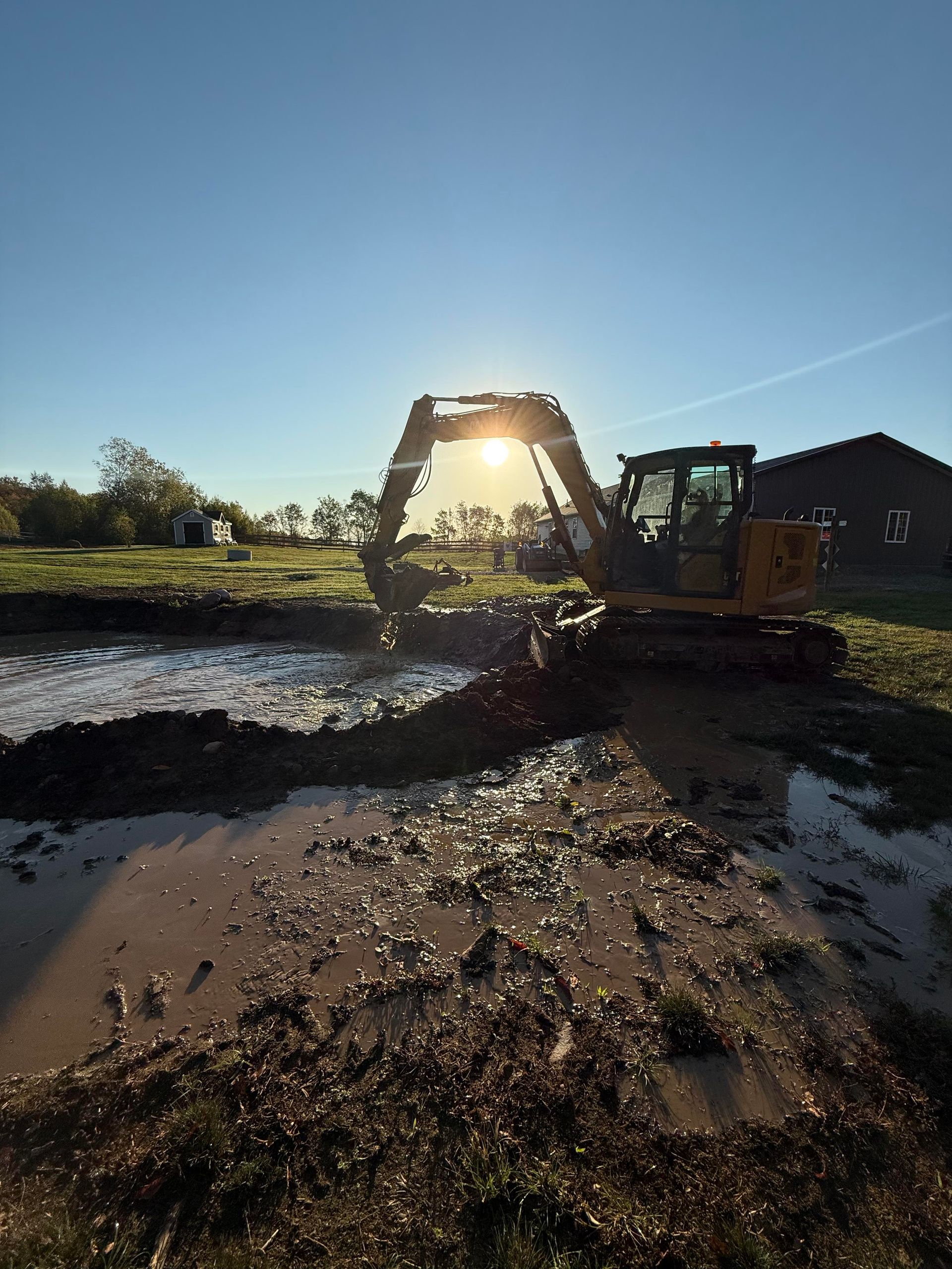 Excavator digging in muddy ground with the sun shining through its arm.