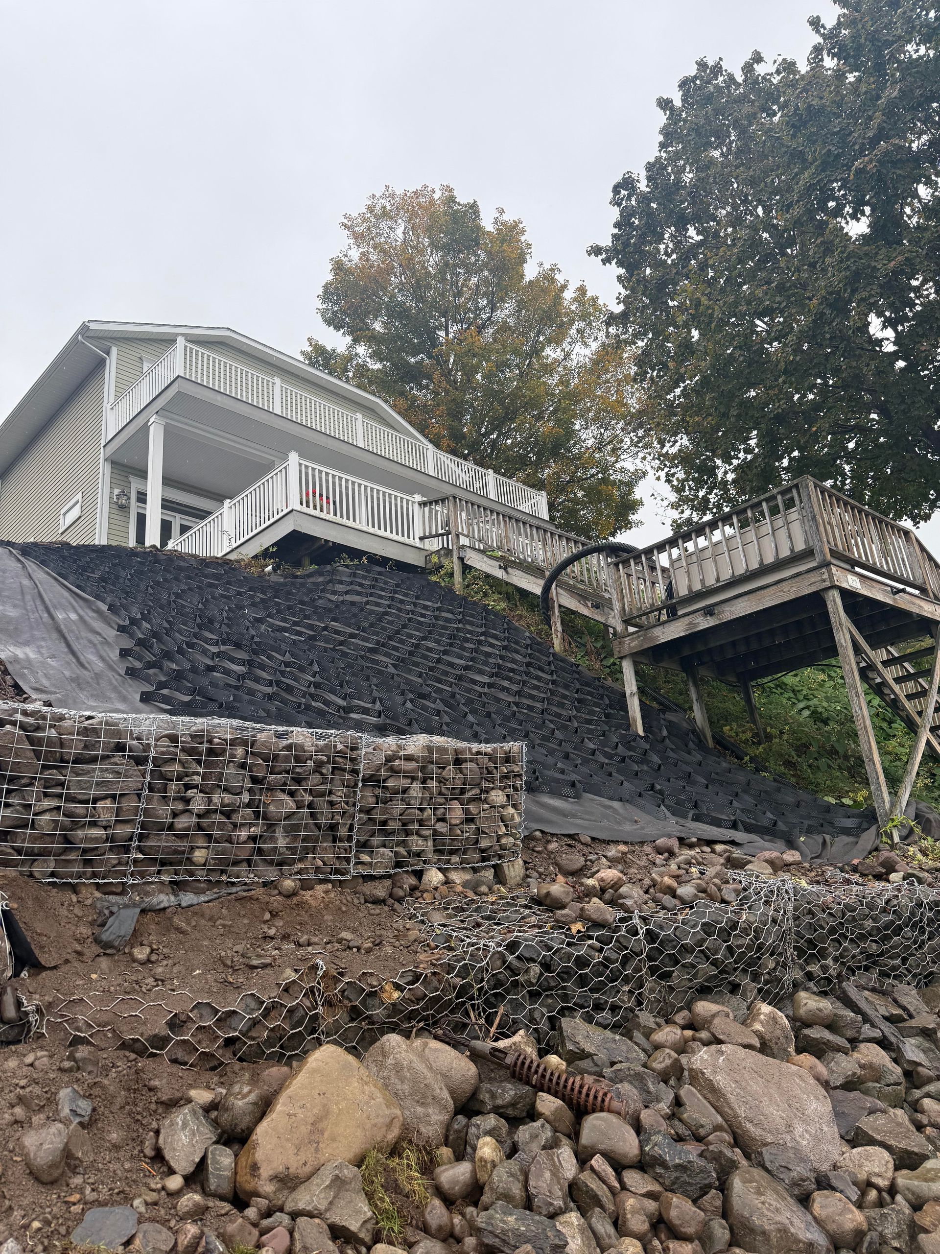 A two-story house with decks on a slope, stabilized by rocks and black gravel. A wooden pier extends right.