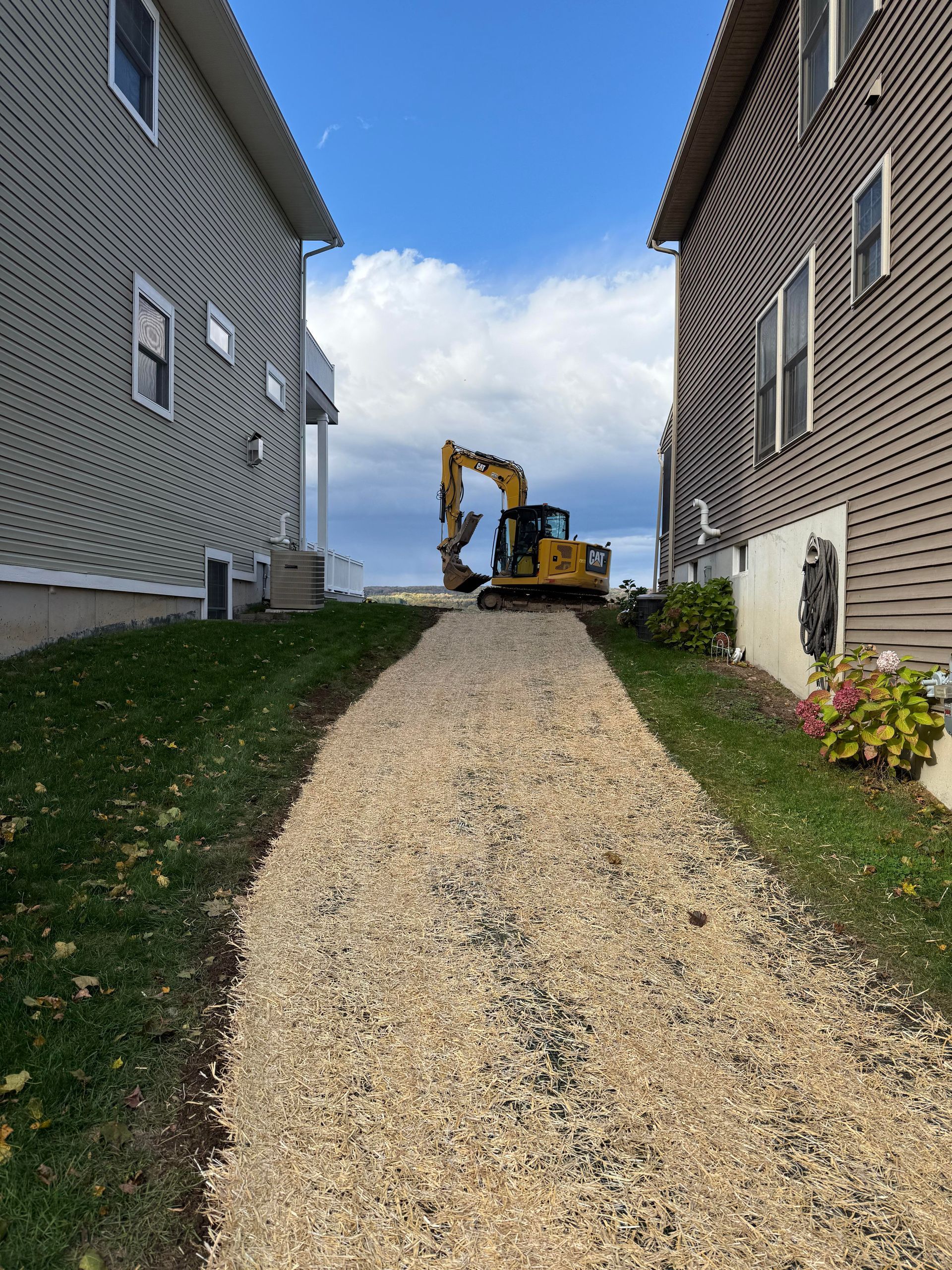 Excavator on gravel path between two houses under a blue sky.