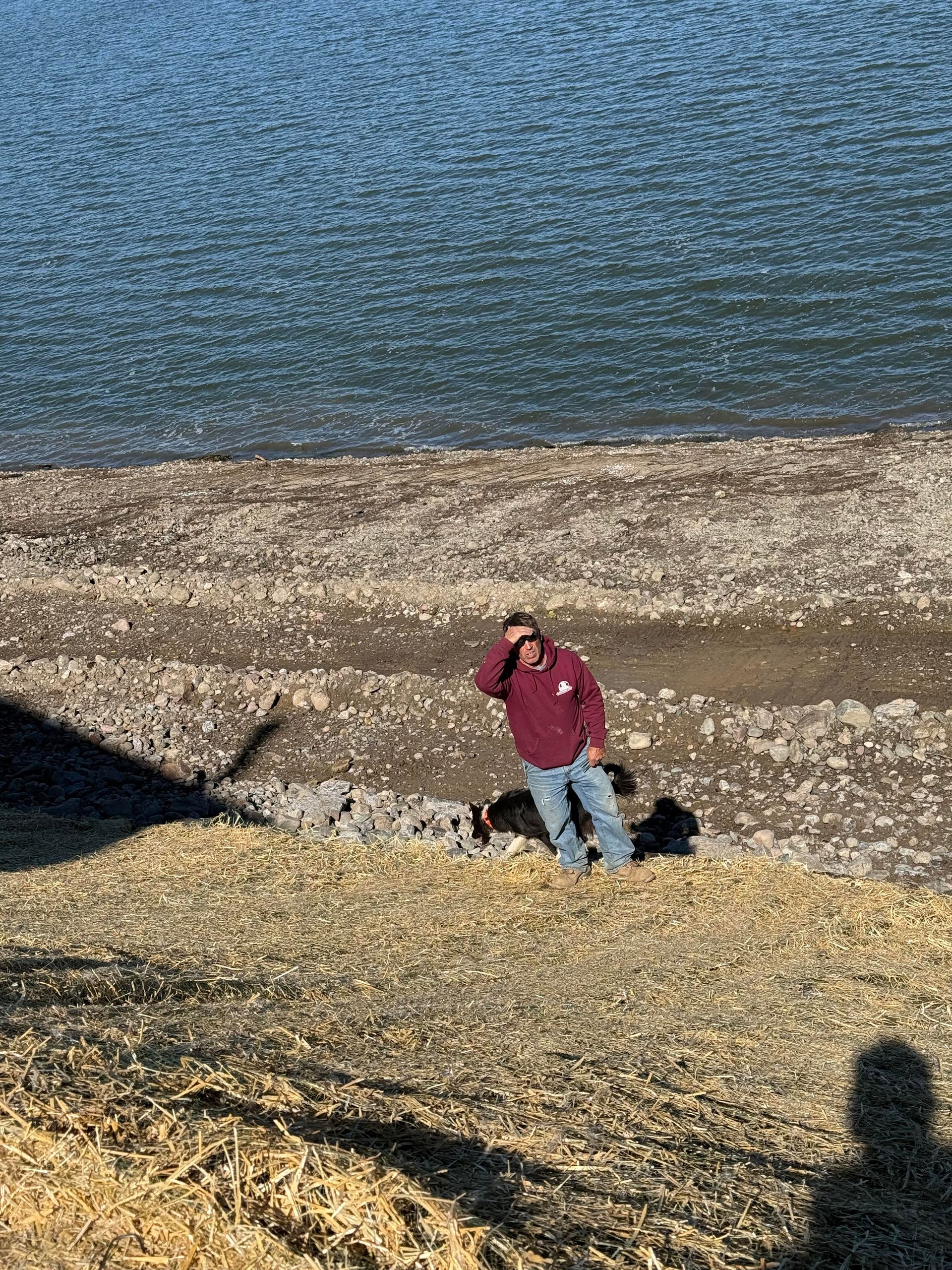 Man in maroon hoodie and jeans stands on a rocky shore, shading eyes, with a body of water in the background.