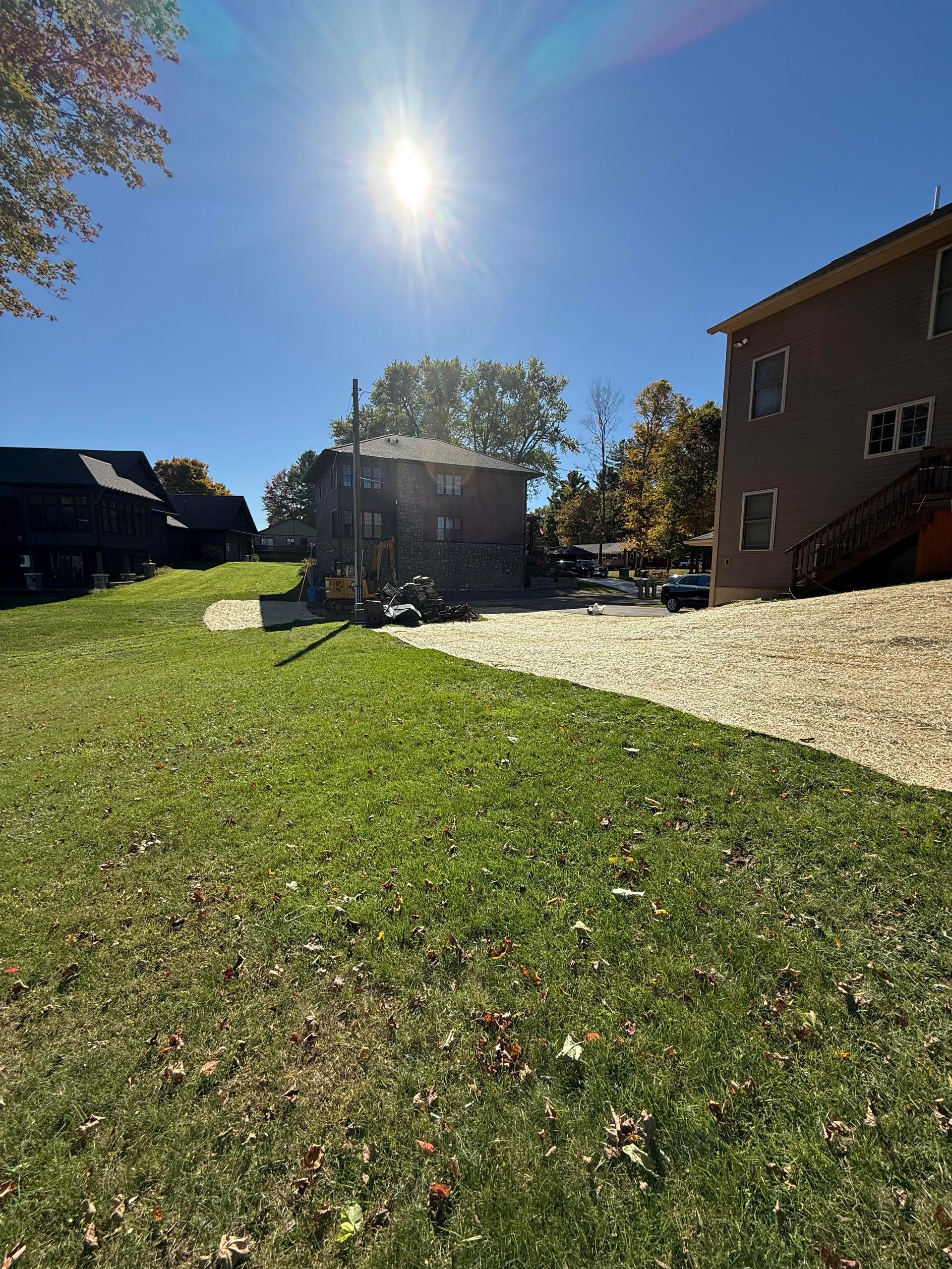 Bright sun over a grassy yard, a gravel path, and two buildings under a clear blue sky.