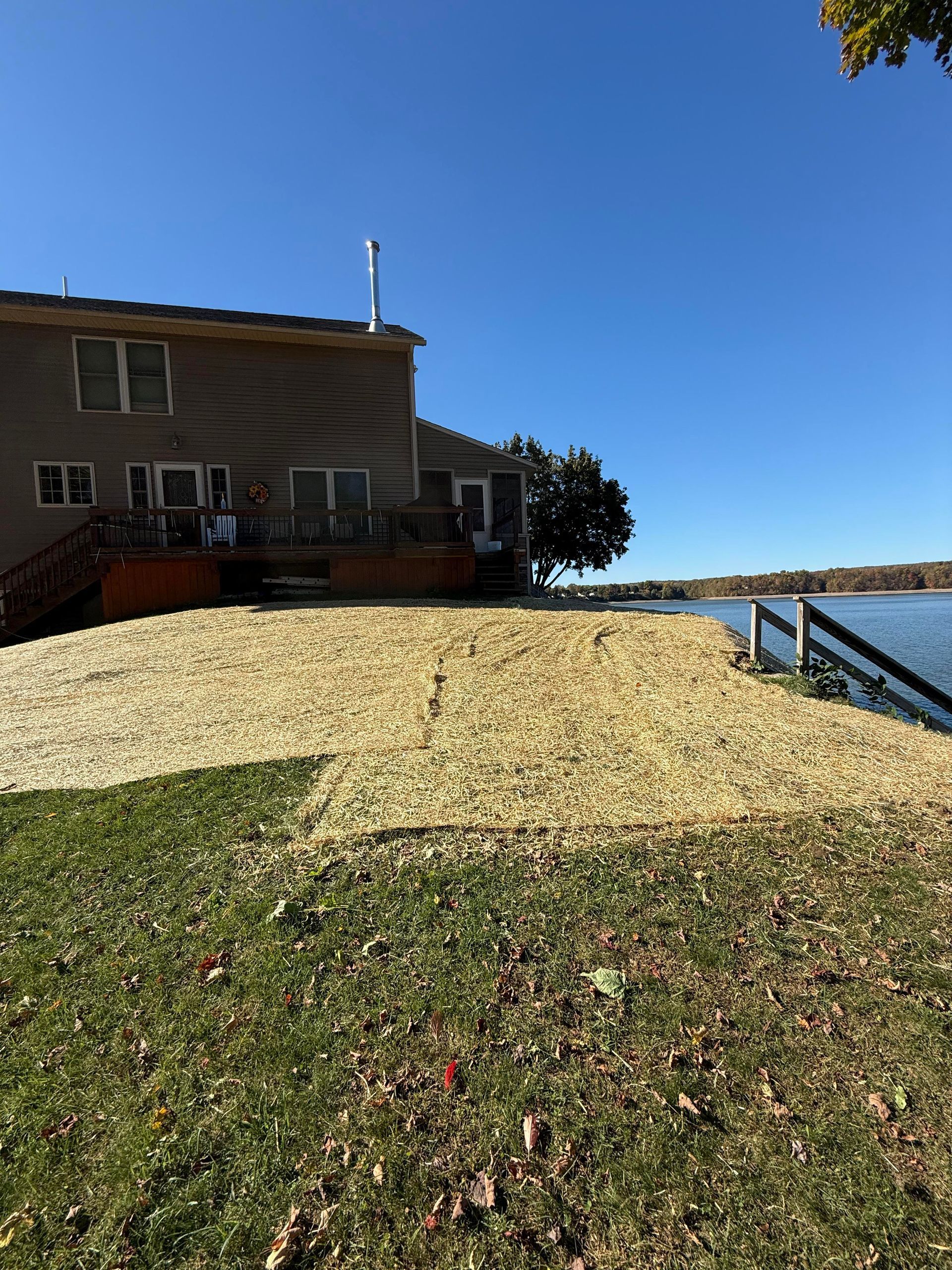 Back of a beige house with a wooden deck overlooking a lake, covered with tan mulch. Blue sky.