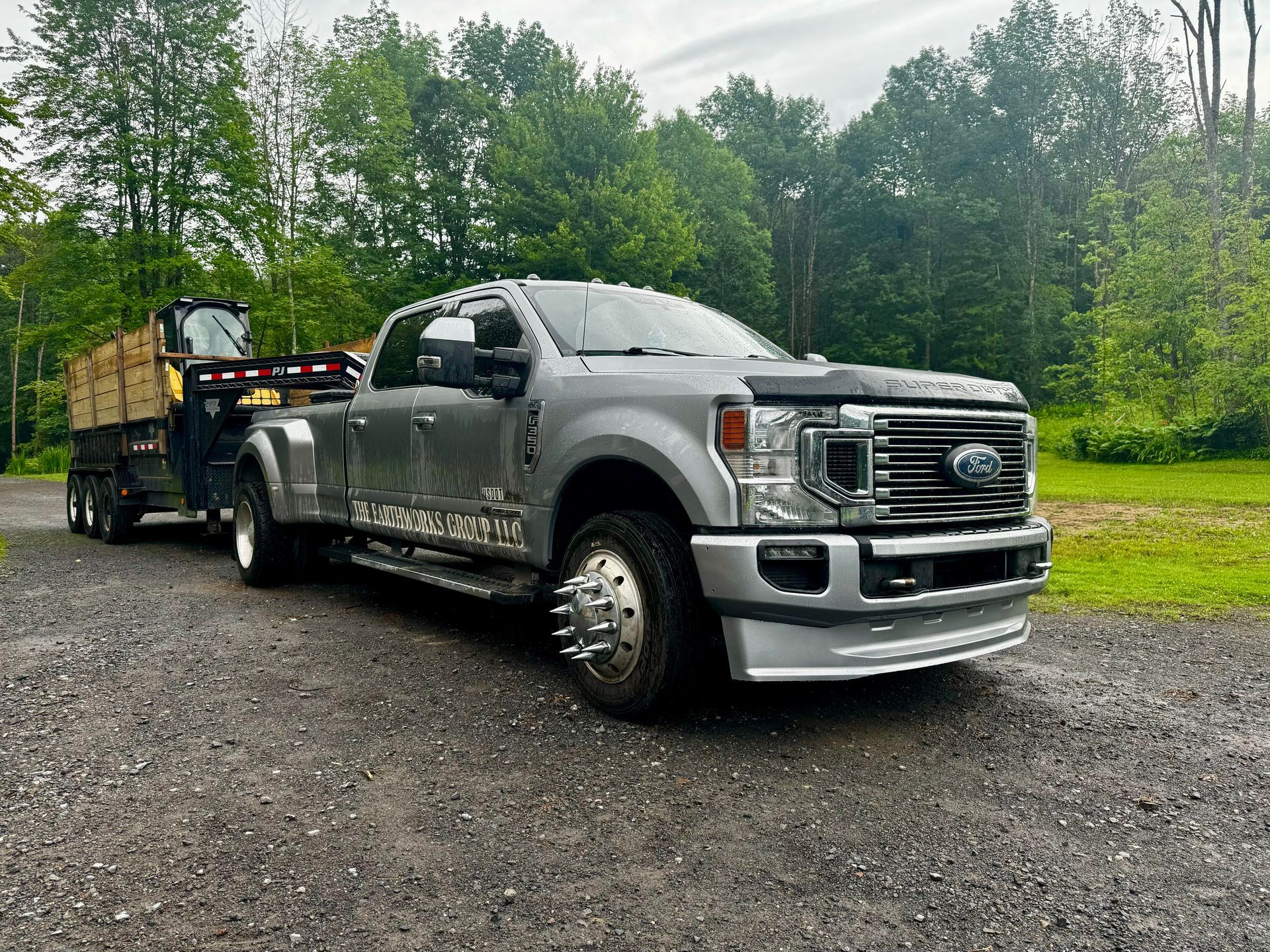 Gray Ford truck towing a trailer with a small excavator on a gravel driveway, trees in background.