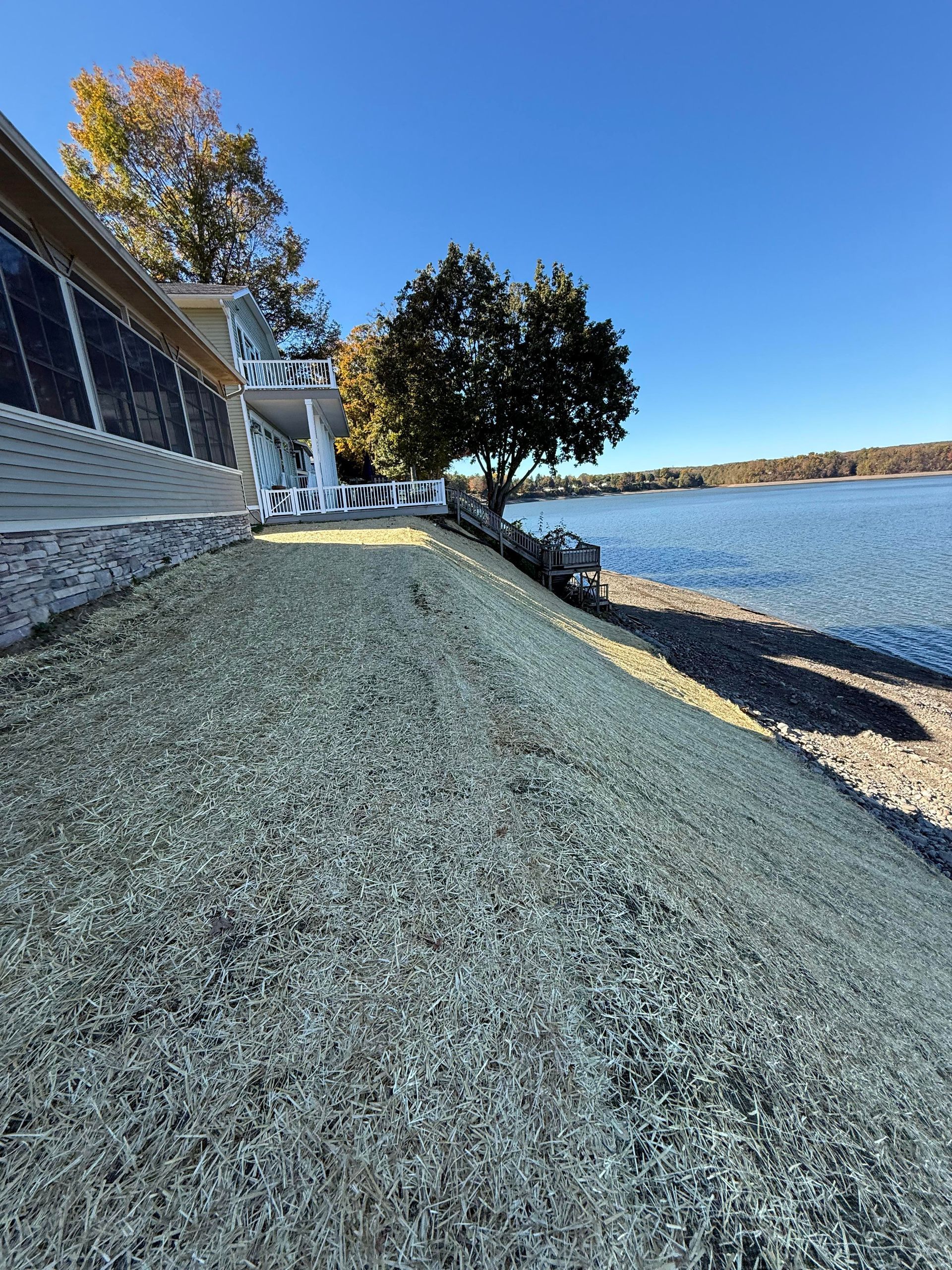 Lakeside house with gravel ground, stone wall, and tree under a clear blue sky.