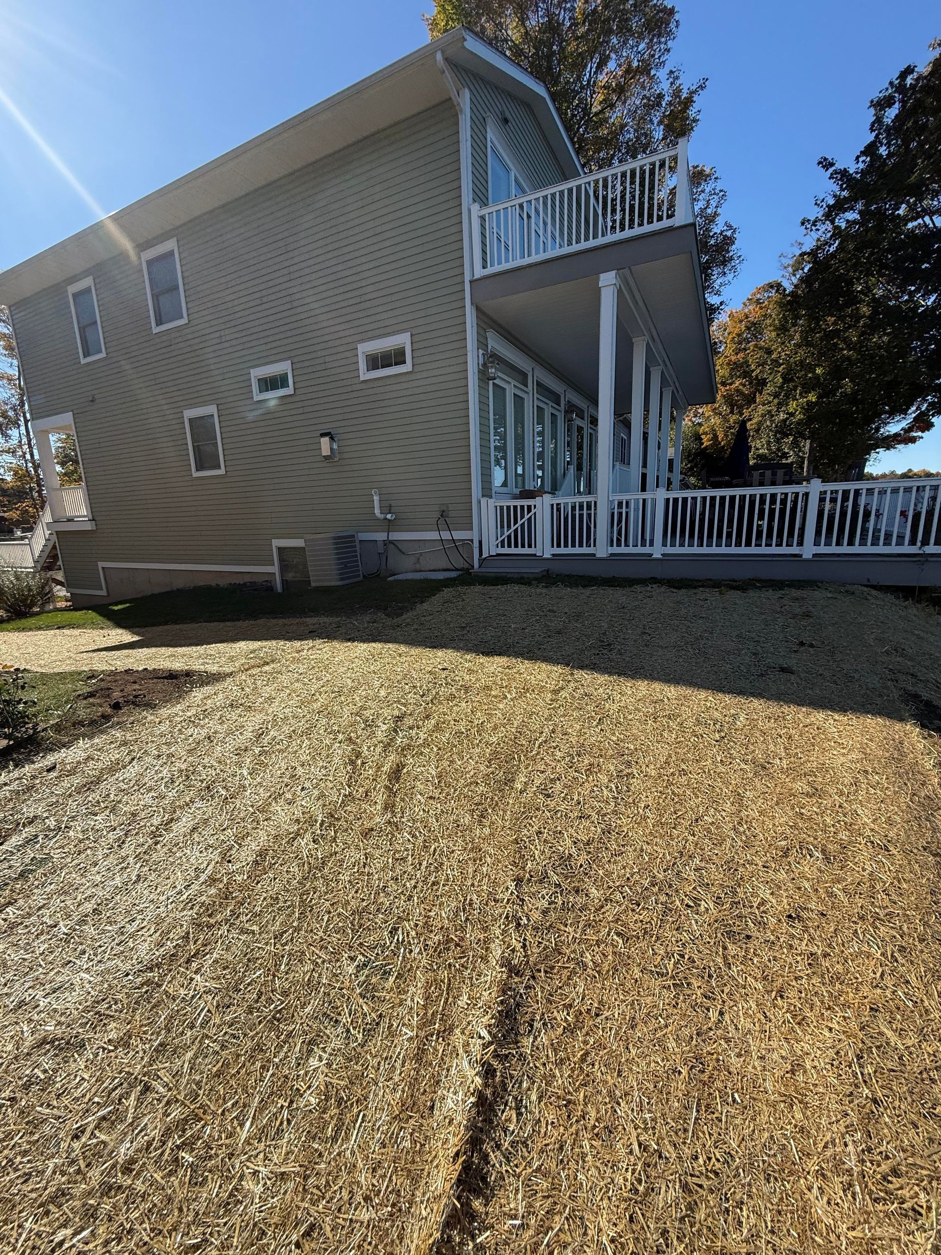 Beige two-story house with a white deck, surrounded by wood chips under a blue sky.
