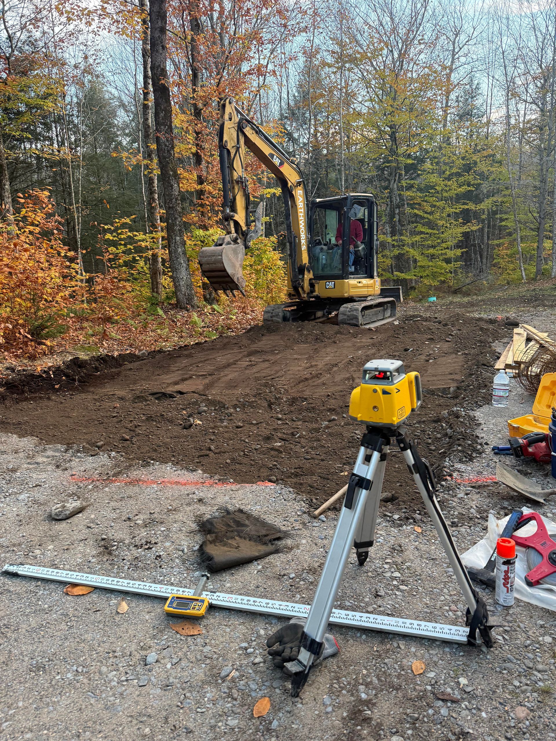 Excavator digging in dirt, a yellow laser level on tripod, trees in background.