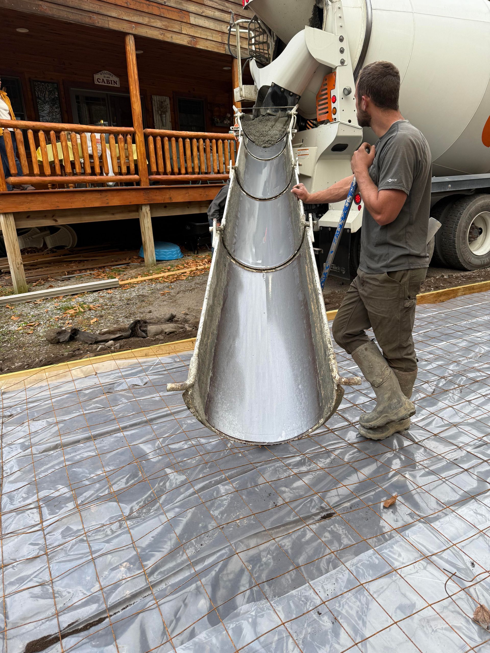 Concrete being poured from a truck chute into a form over rebar and plastic sheeting, worker guiding.