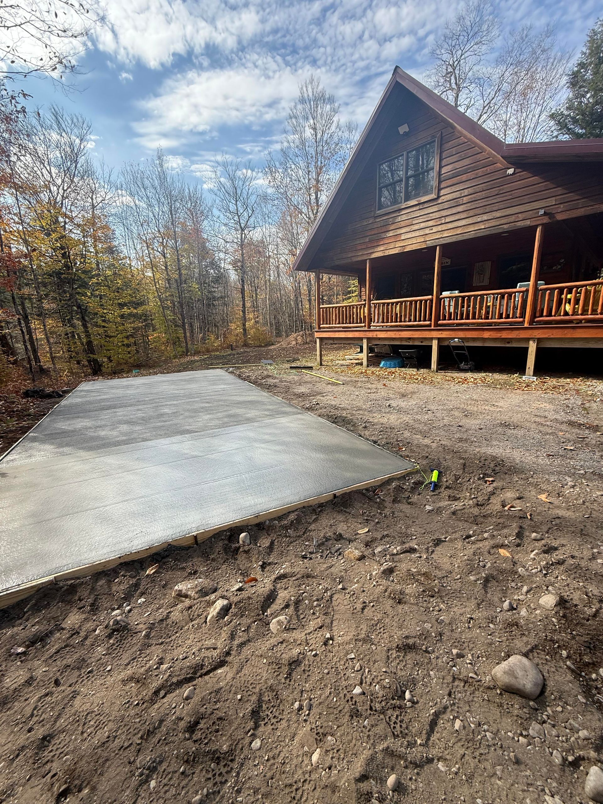 Concrete pad in front of a wood cabin, surrounded by dirt and trees, under a cloudy sky.